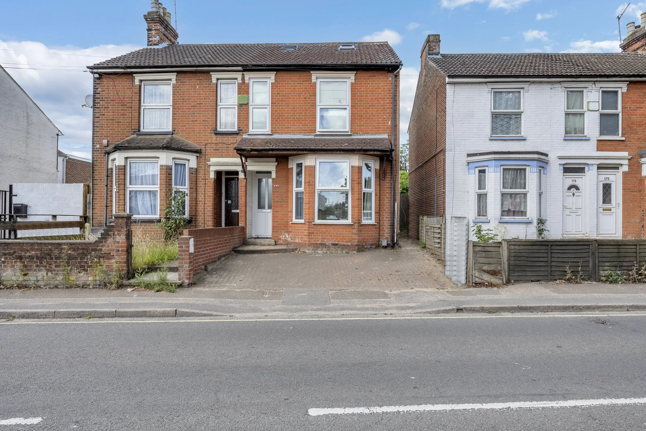 A row of houses along a street with a brick house in the center, a driveway in front of it, and neighboring houses on each side, under a blue sky with clouds.