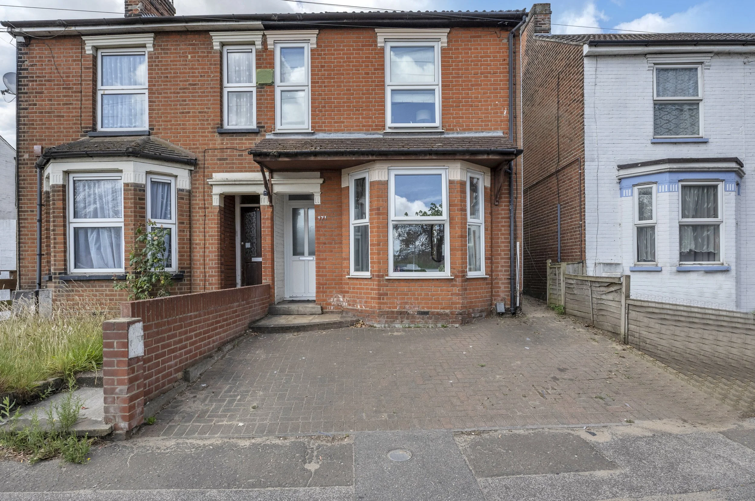 A red brick two-story house with a small front step, white door, and large front window, situated between two other houses, one blue and one white, on a quiet street with a paved driveway.