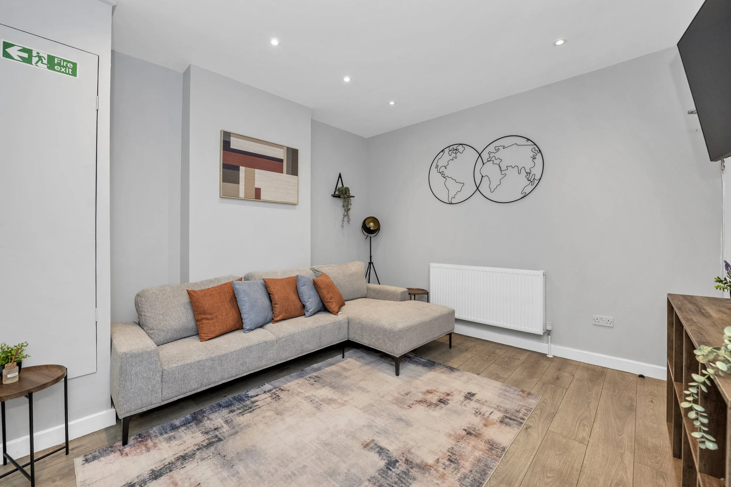 Modern living room with a beige sectional sofa with orange and blue pillows, a world map wall decor, a floor lamp, a wooden side table, a rug, and framed artwork on the wall.