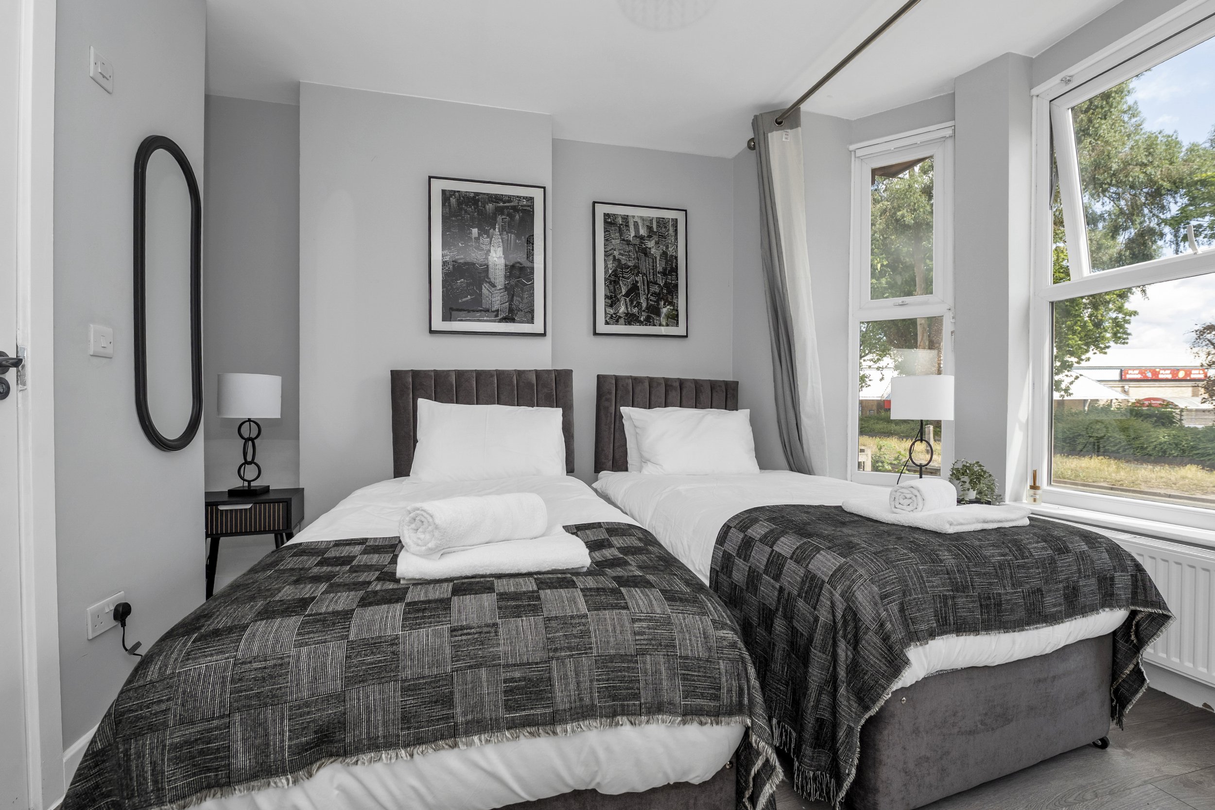 A modern bedroom with two twin beds, each with a brown upholstered headboard, white bedding, and a rolled towel at the foot. There are two black nightstands with white lamps, black and white framed cityscape art on the wall, and large windows with white curtains letting in natural light.