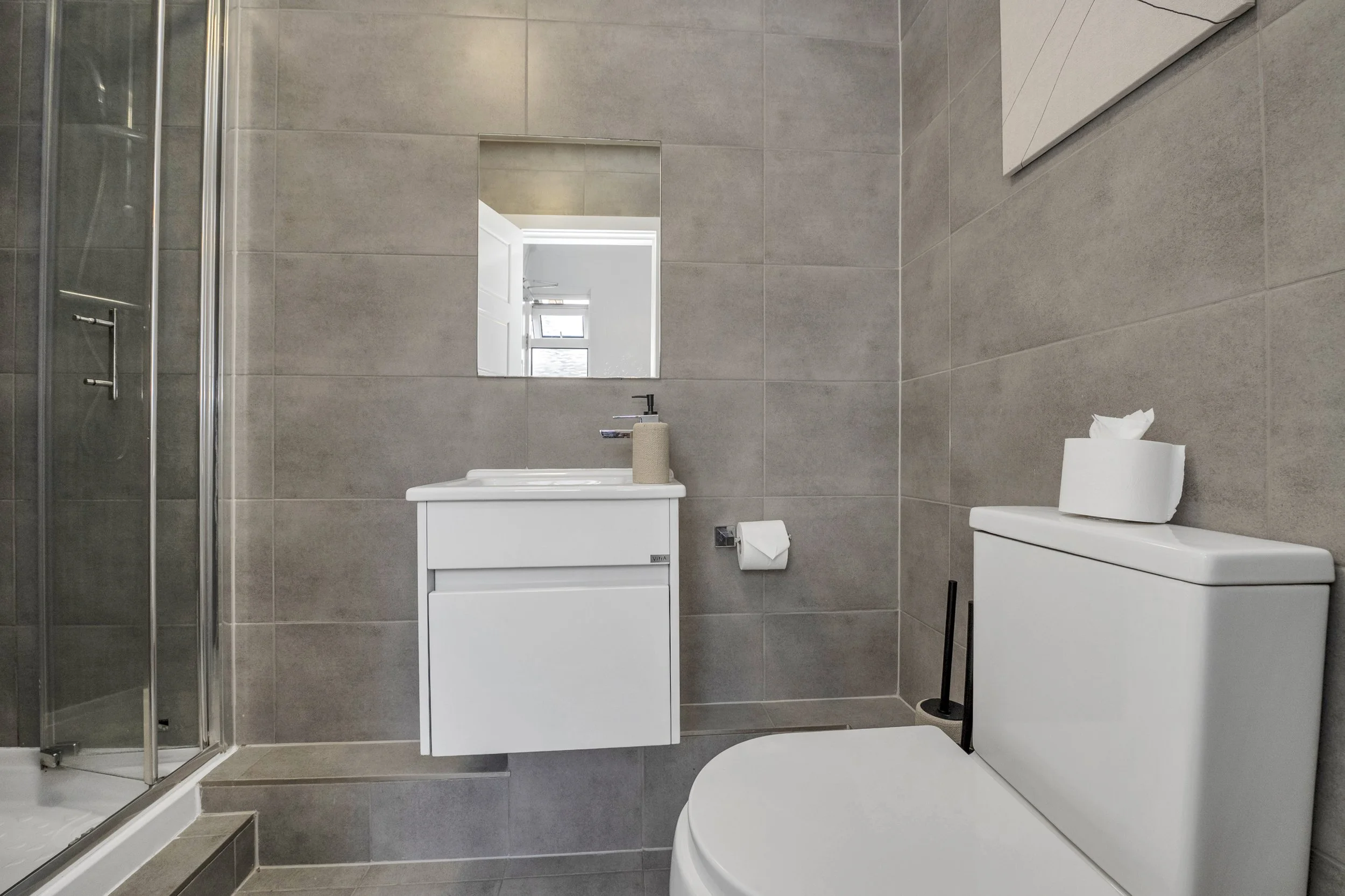 Modern bathroom with gray tiled walls, a white toilet, a small white vanity with a mirror, a paper towel roll, and a shower enclosure in the corner.
