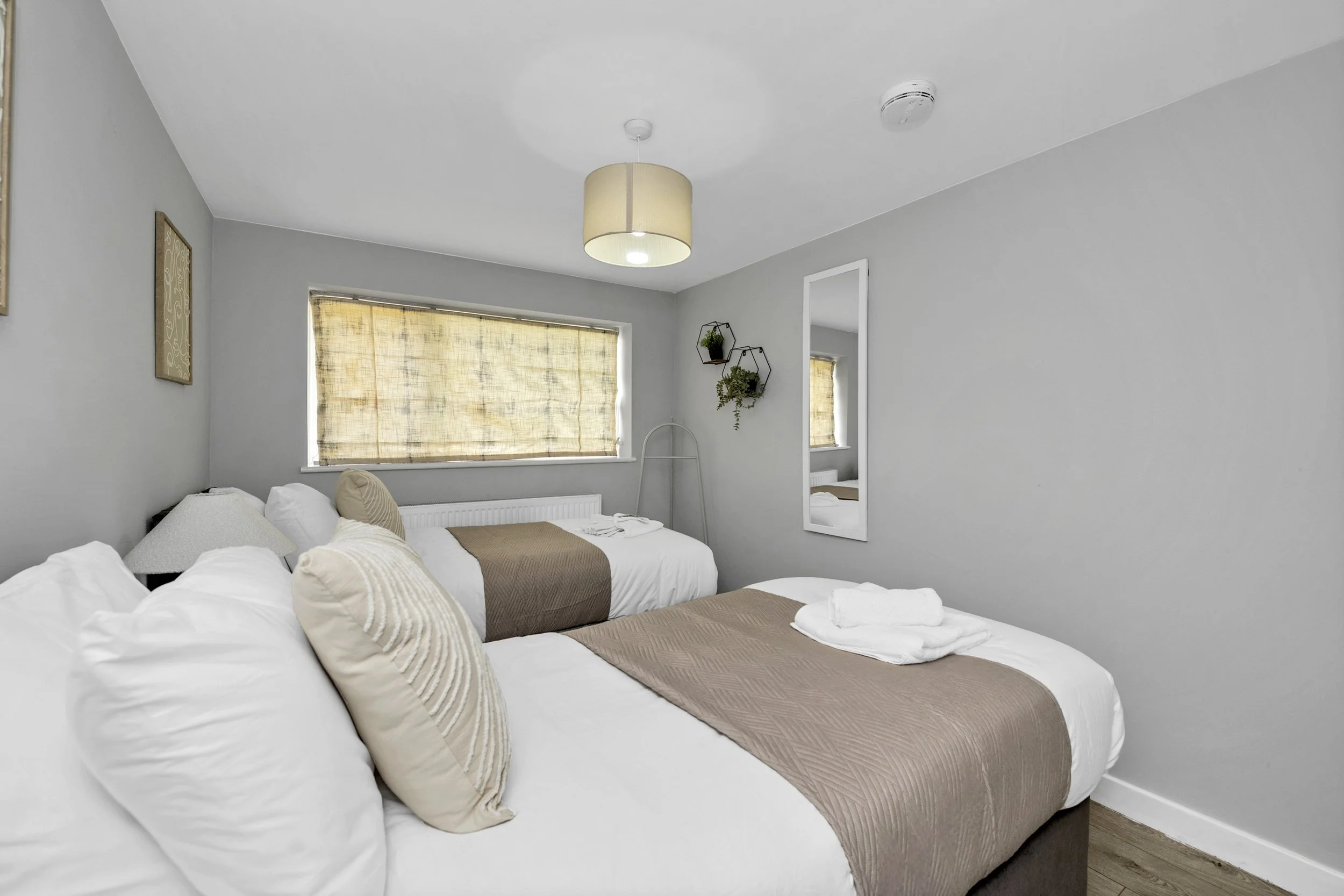 A clean, minimalist bedroom with two beds, beige bedding, a window with a beige shade, and wall decorations, including a mirror and hanging plants.