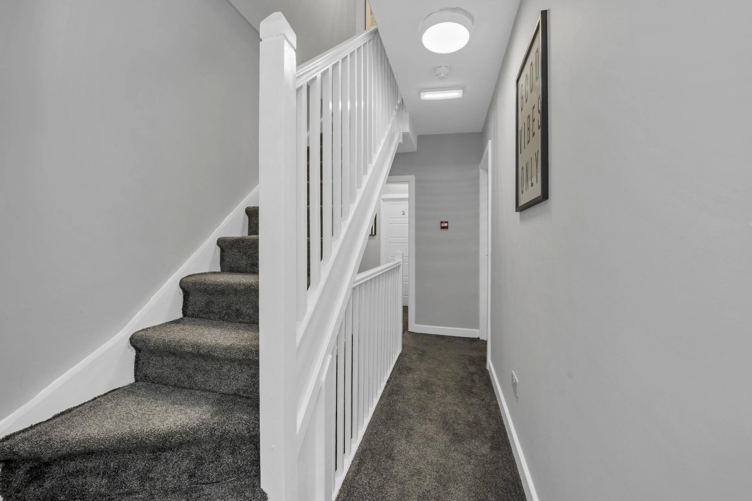A hallway with gray walls, dark carpet, white staircase with a railing, and a framed wall art piece. Ceiling lights and a door at the end of the hall are visible.