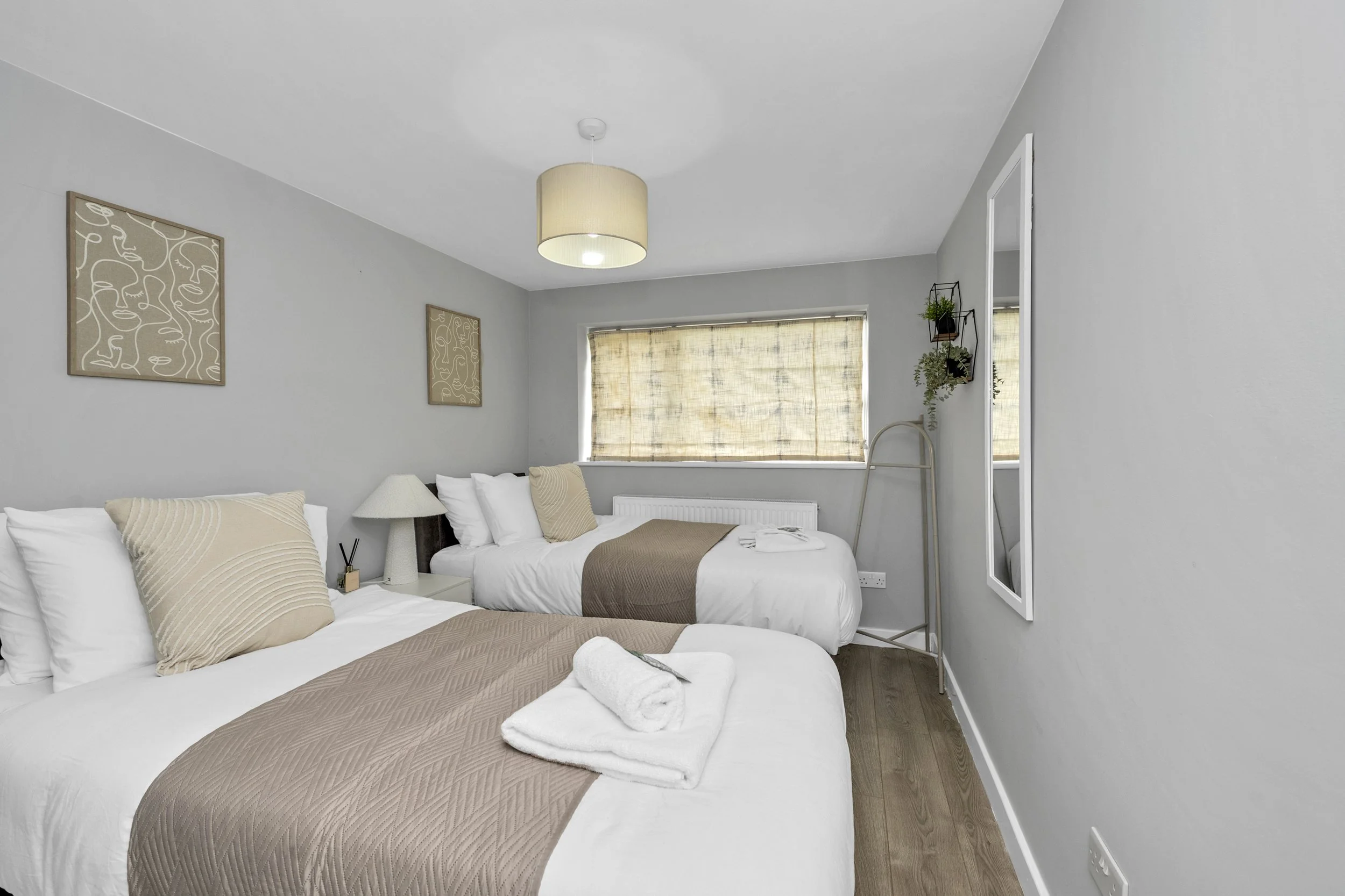 A tidy bedroom with two white beds, beige and white pillows, a beige bed runner, and neatly folded white towels on one bed. The room features a window with beige shades, a standing mirror, wall art, a small lamp, and some potted plants.