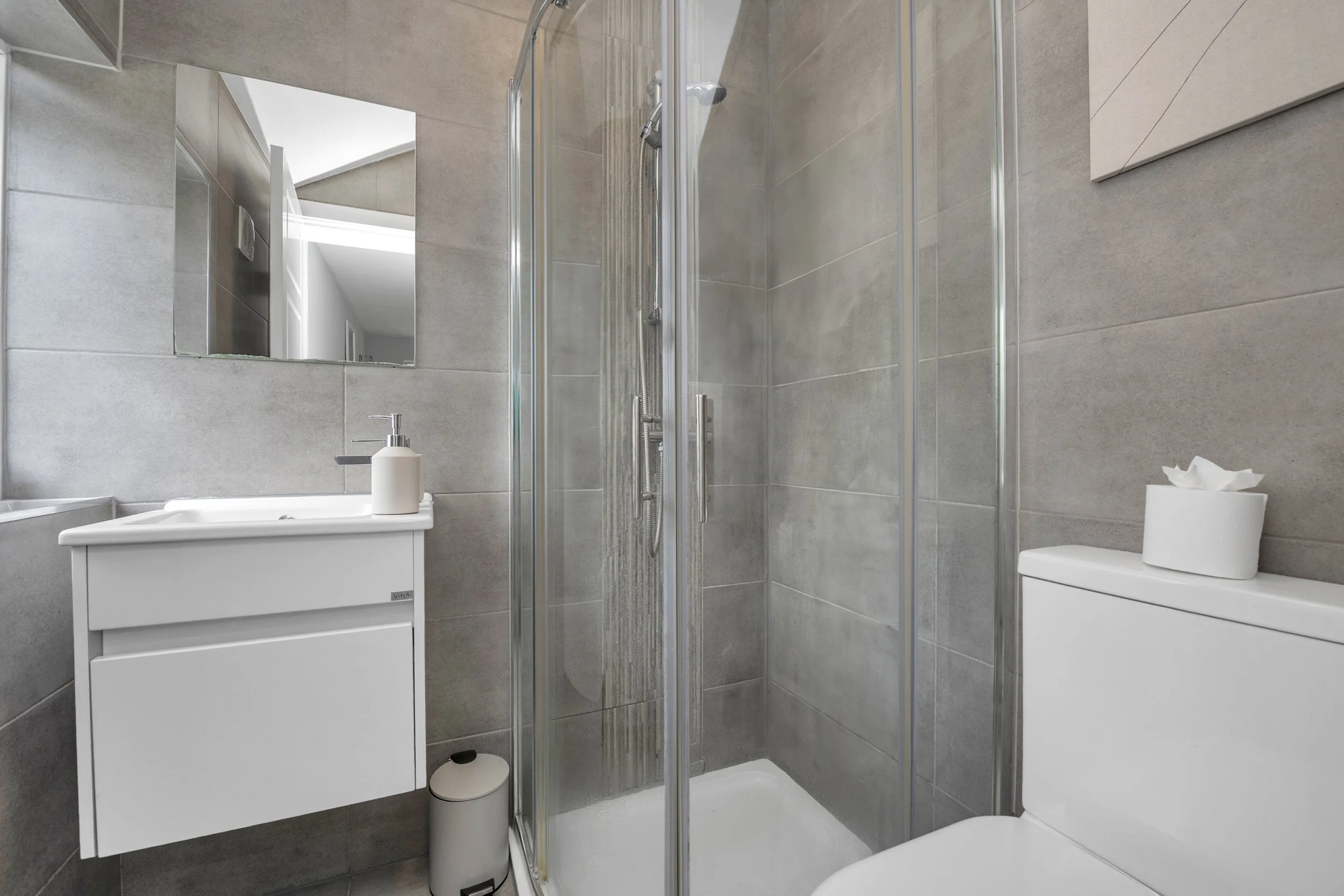 Modern bathroom featuring a glass shower enclosure, a white wall-mounted vanity with a sink and a soap dispenser, a mirror above the vanity, a white toilet with a tissue box on top, and neutral gray tiles on the walls.