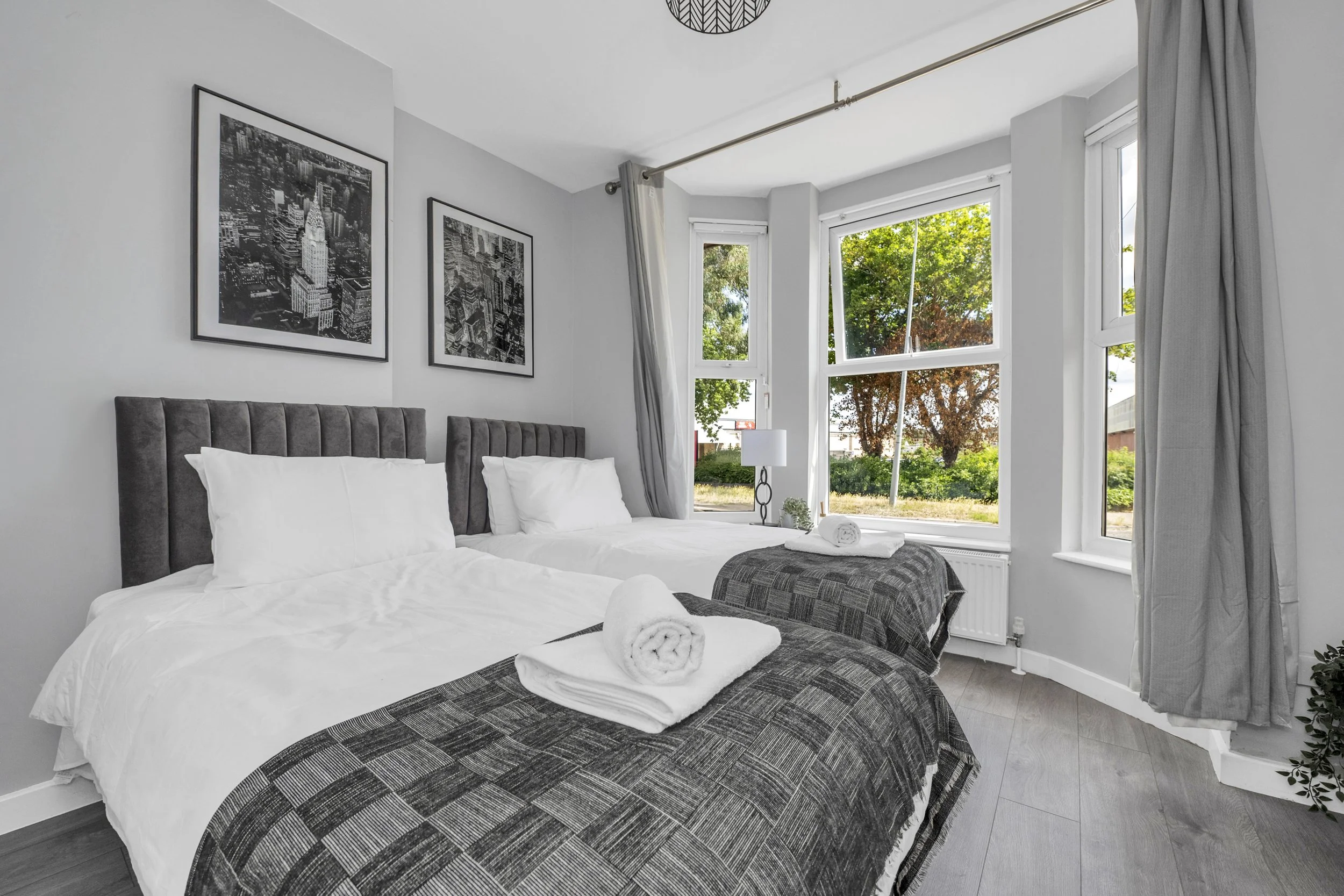 A bright bedroom with two beds, gray headboards, white bedding, and dark gray patterned blankets. Towels are folded on each bed. There are two black-and-white cityscape pictures on the light gray wall. Large windows with gray curtains reveal green trees outside.