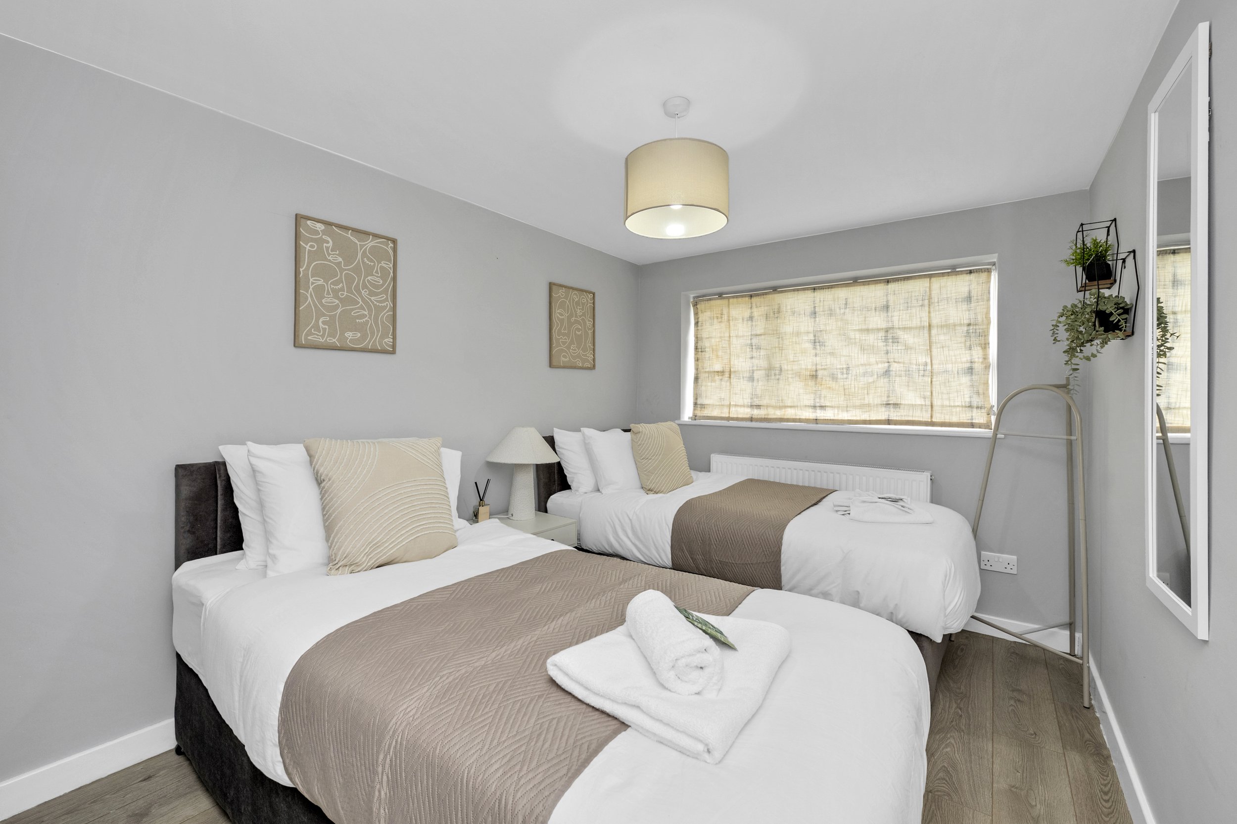 A bedroom with two single beds, beige bedding, pillows, a nightstand with a lamp, and a large window with blinds.