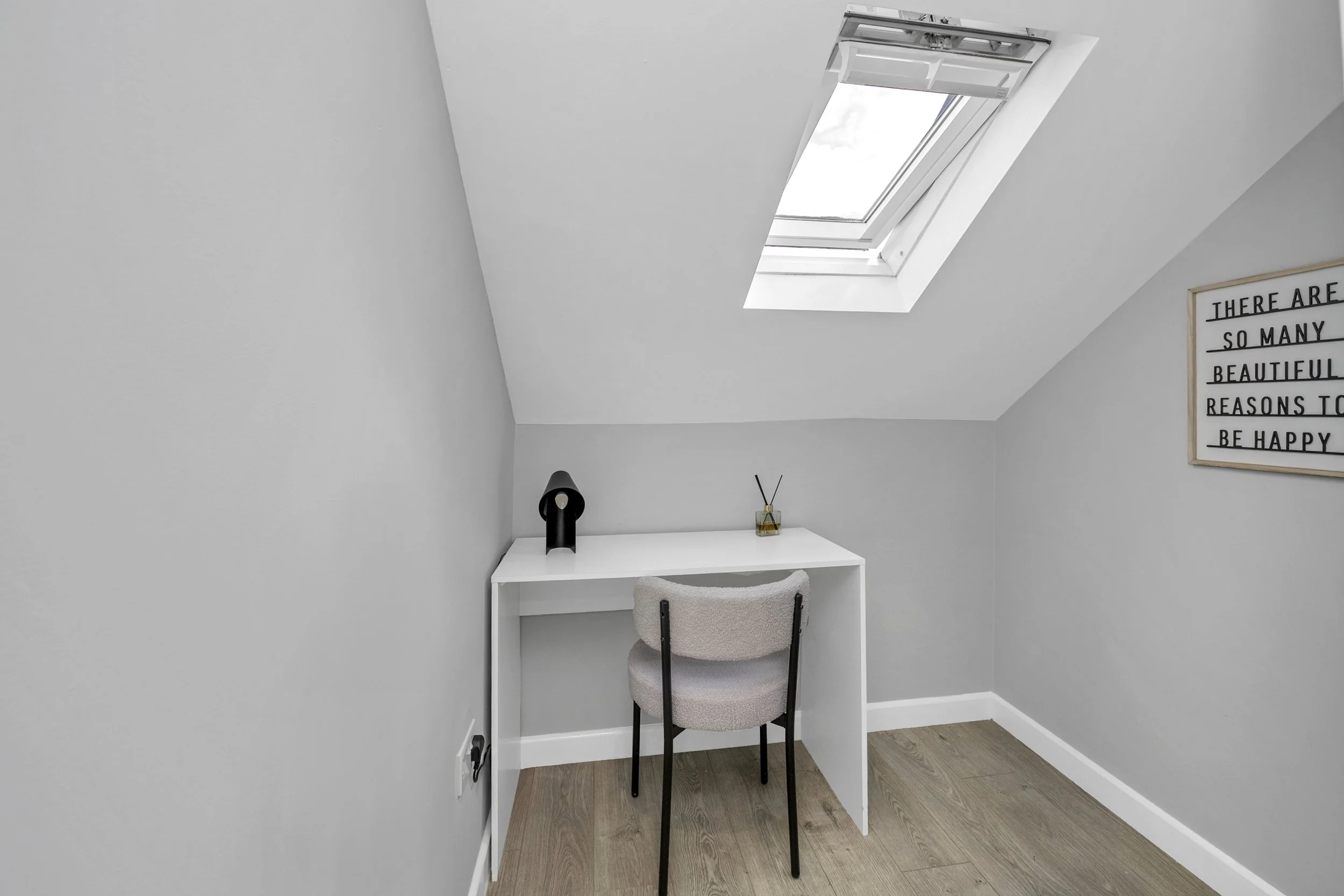 A small minimalist workspace with a white desk, a beige chair, a black modern desk lamp, and a small jar with reed diffusers. The room has gray walls, a wooden floor, and a skylight window on the sloped ceiling. A framed quote on the wall reads 'There are so many beautiful reasons to be happy.'