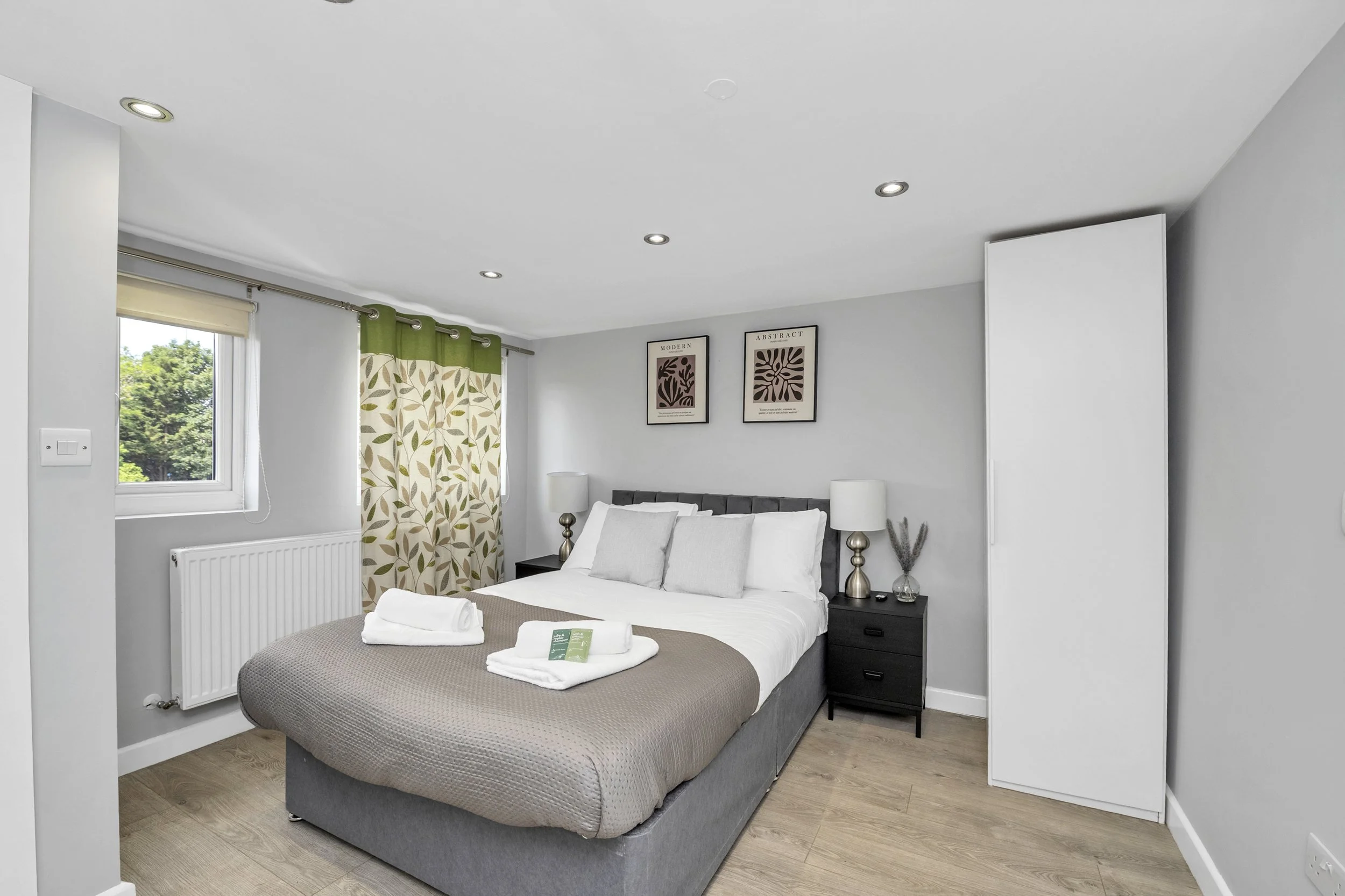 A neatly made bed in a modern bedroom with two bedside tables, lamps, framed wall art, window with patterned curtains, and a white wardrobe in the corner.