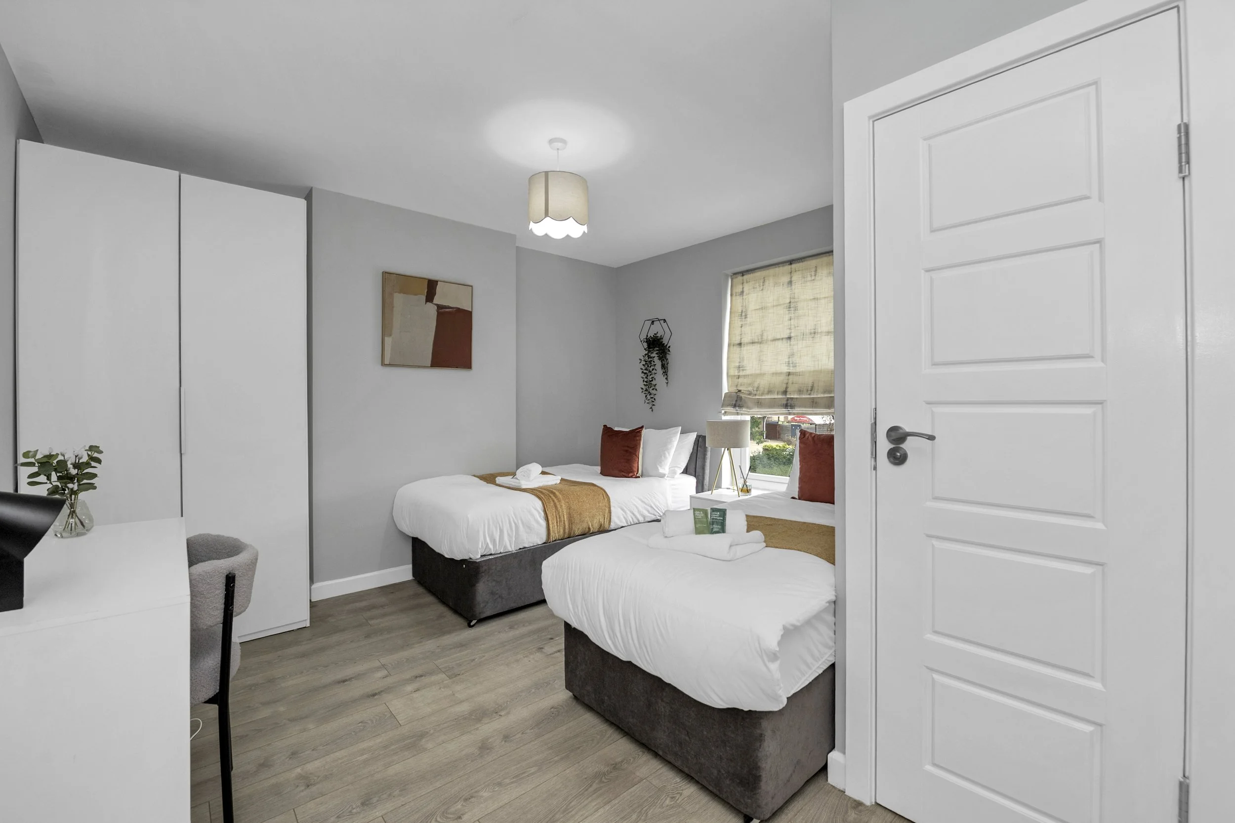 Two single beds in a modern bedroom with white linens, brown and white pillows, and a mustard-colored throw blanket. There is a window with a beige Roman shade, a wall-mounted plant, a bedside table with a lamp, and a small vanity or desk with a chair. The room has gray walls, light wood flooring, and minimalistic decor.