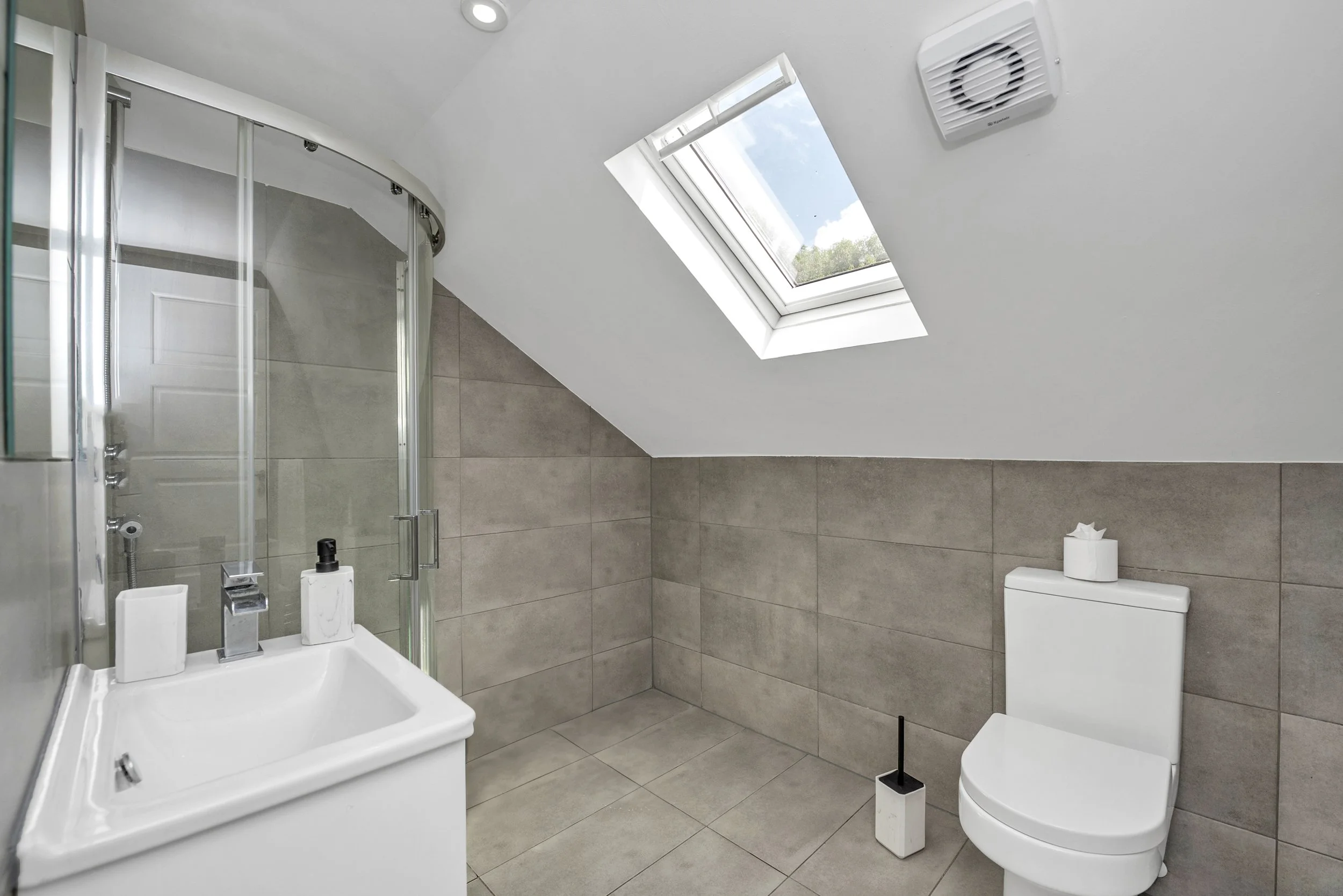 Modern bathroom with skylight, shower enclosure, white sink, toilet with tissue on top, and neutral-colored tiles.