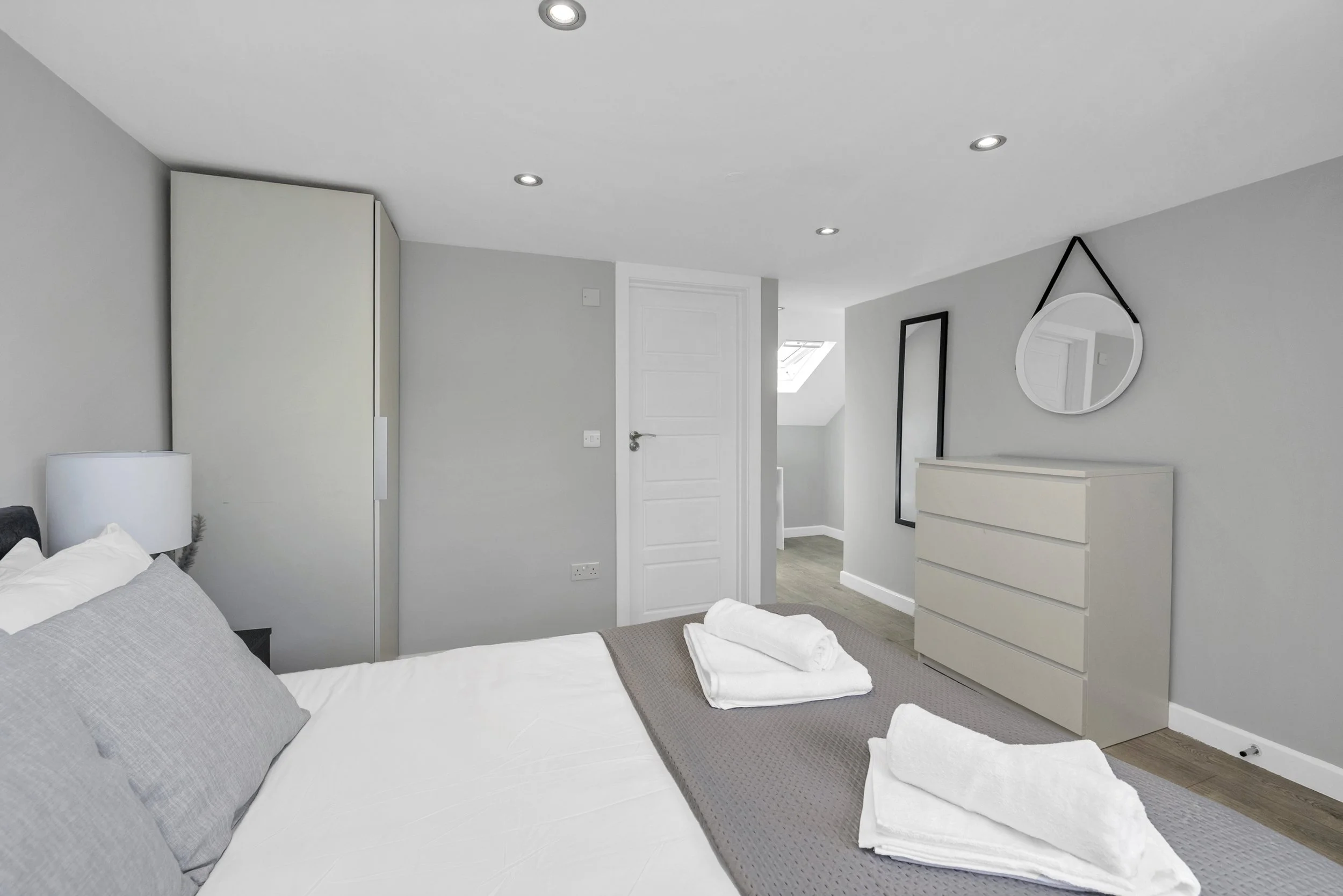 A bedroom with a bed, gray pillows, and white linens, with two folded towels on the bed. There is a white nightstand and a white dresser on the wall. The walls are painted light gray, and there are three recessed ceiling lights. A mirror is hanging on the wall, and there is a door and small hallway visible.