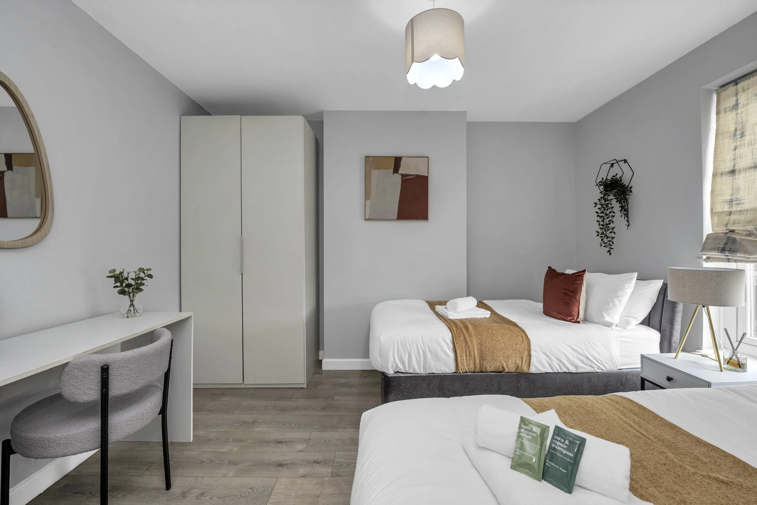 A modern bedroom with two beds, a white desk with a grey chair, a wardrobe, a wall mirror, and decorative plants. Natural light from a window with a beige curtain.