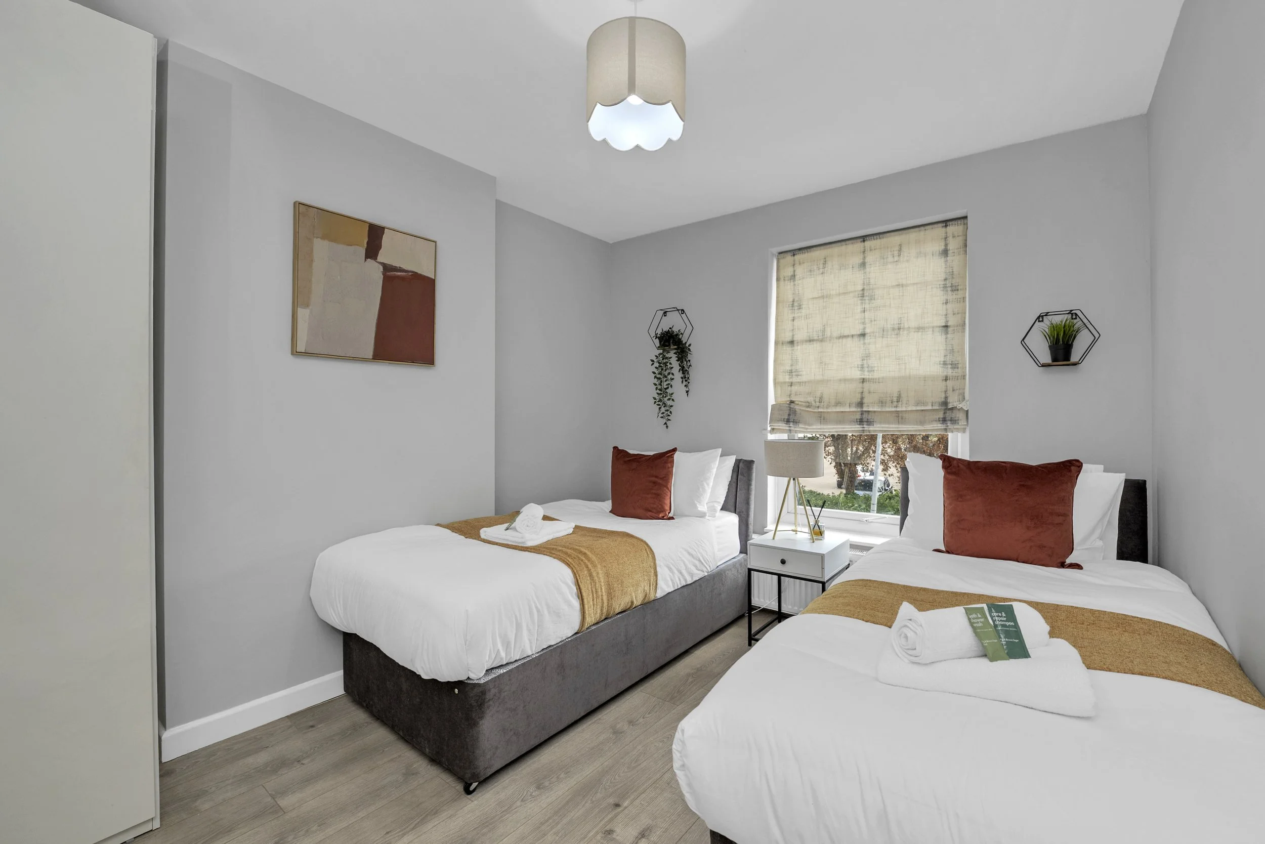 A bedroom with two single beds, each with white linens and rust-colored pillows. A white nightstand with a lamp sits between the beds, and a window with a beige Roman shade is behind the beds. There are two small wall shelves with plants and a piece of abstract artwork on the wall.