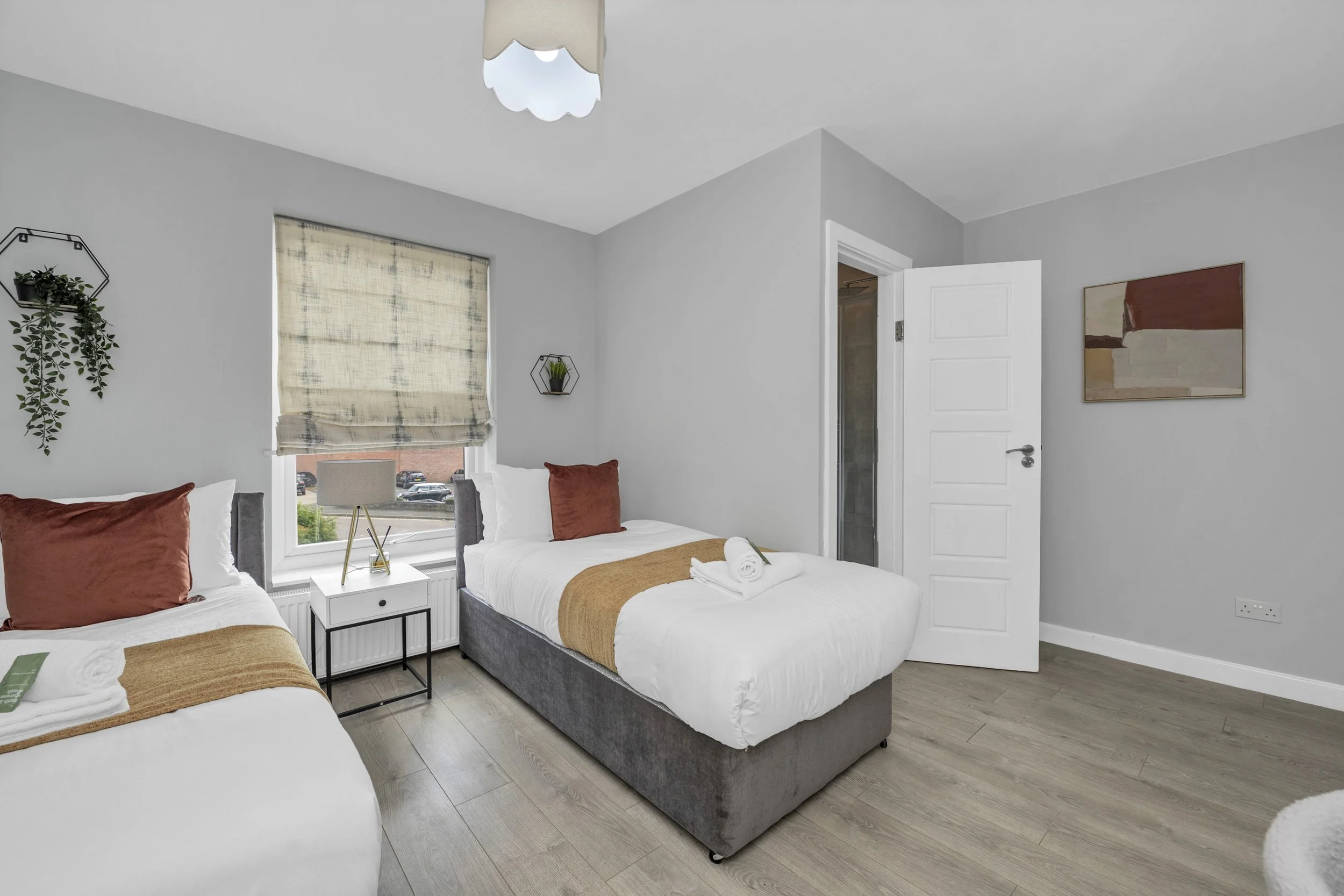 A modern bedroom with two beds, each with white bedding and rust-colored pillows, a window with a beige blind, a bedside table with a lamp, and contemporary wall decorations.