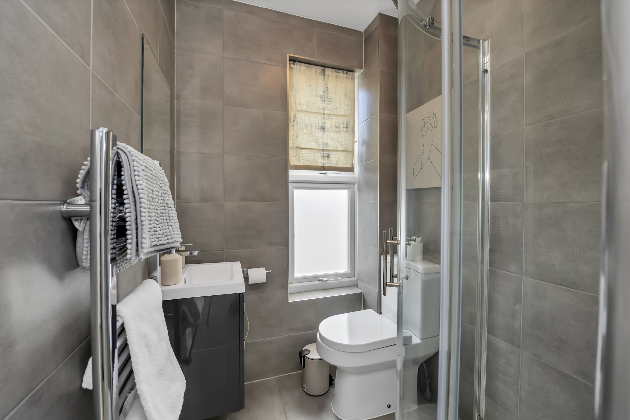 Modern bathroom with gray tiles, a small sink, a toilet, a frosted window, and a glass shower enclosure.