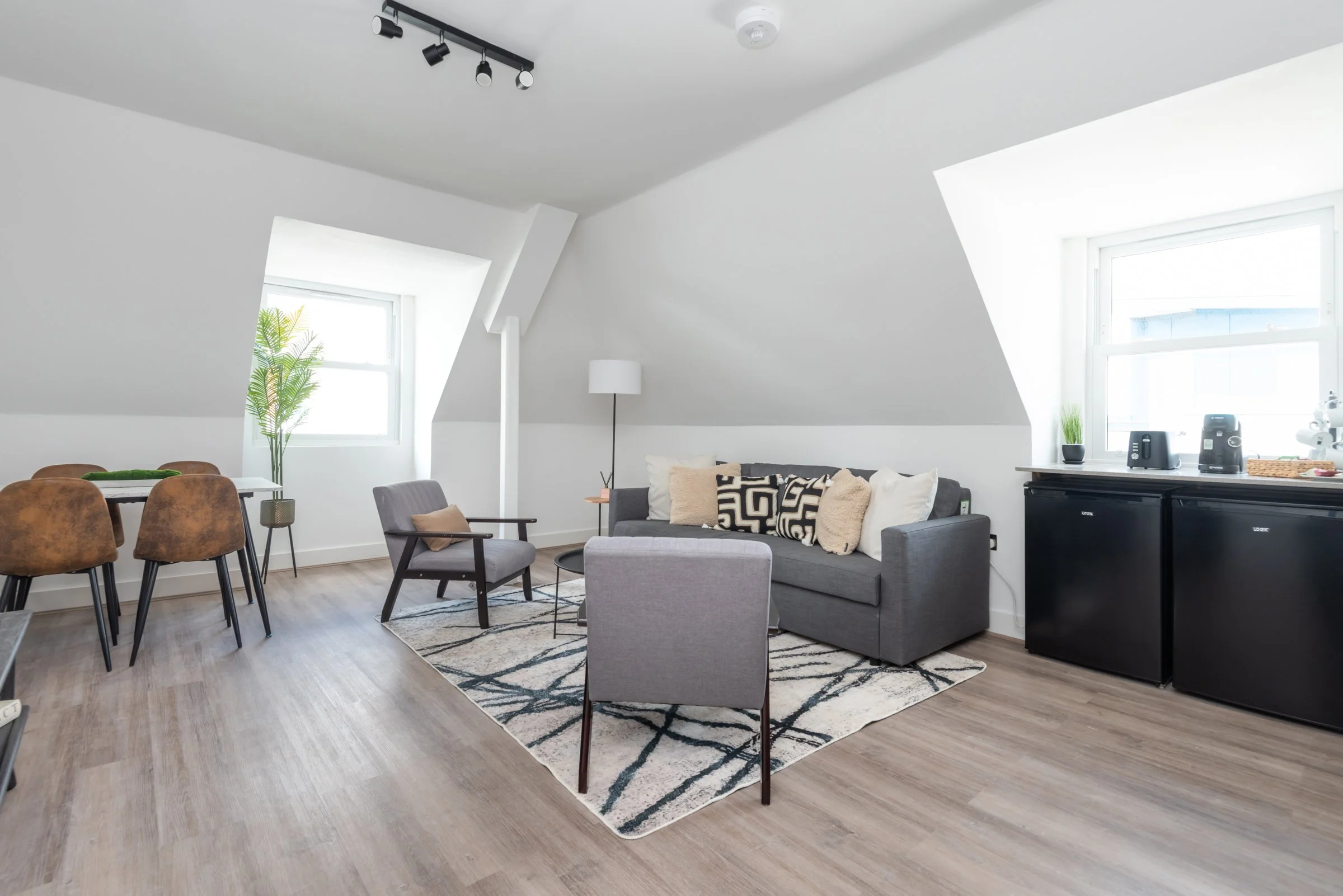 Modern living room in an attic with white walls, a gray sofa, and a patterned rug, surrounded by gray and brown chairs, with windows on sloped ceilings.