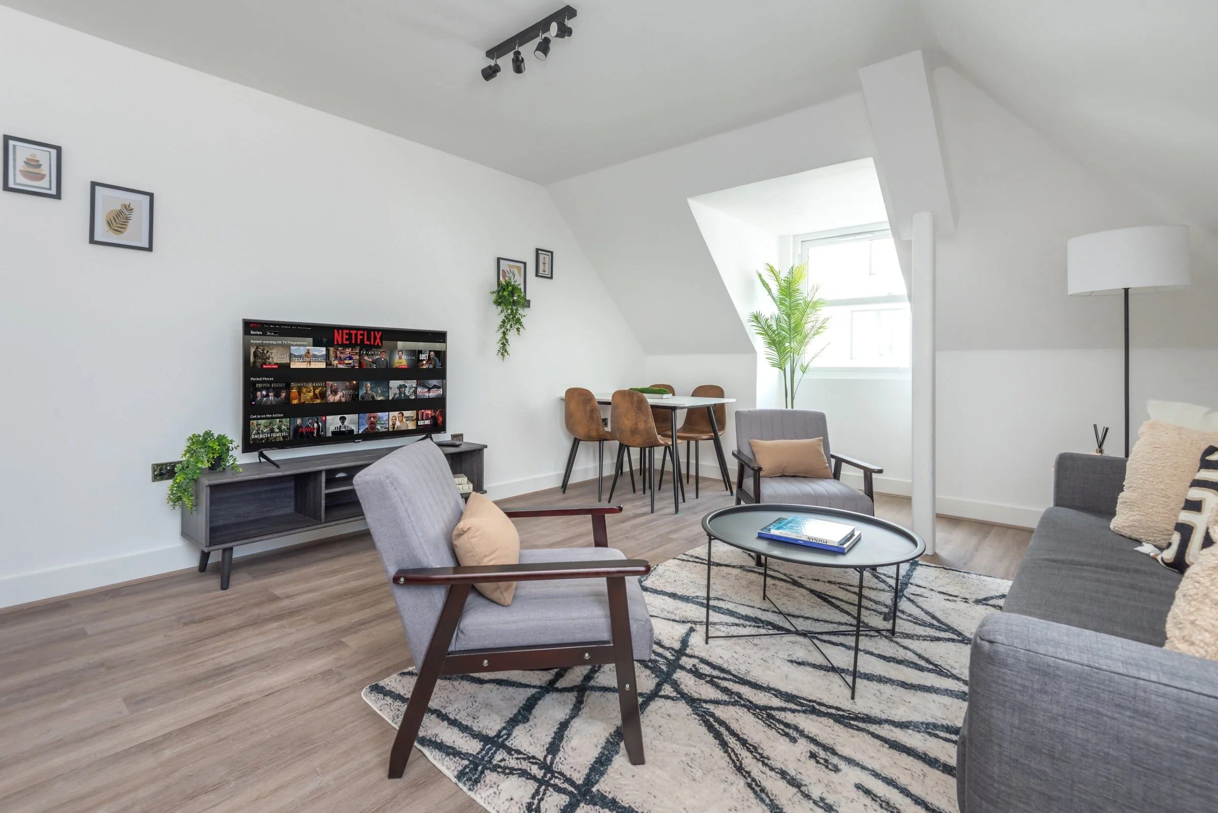 Modern living room with gray sofas, wooden chairs, a black coffee table, a media console with a TV showing Netflix, potted plants, framed artwork on the wall, and a large window bringing in natural light.