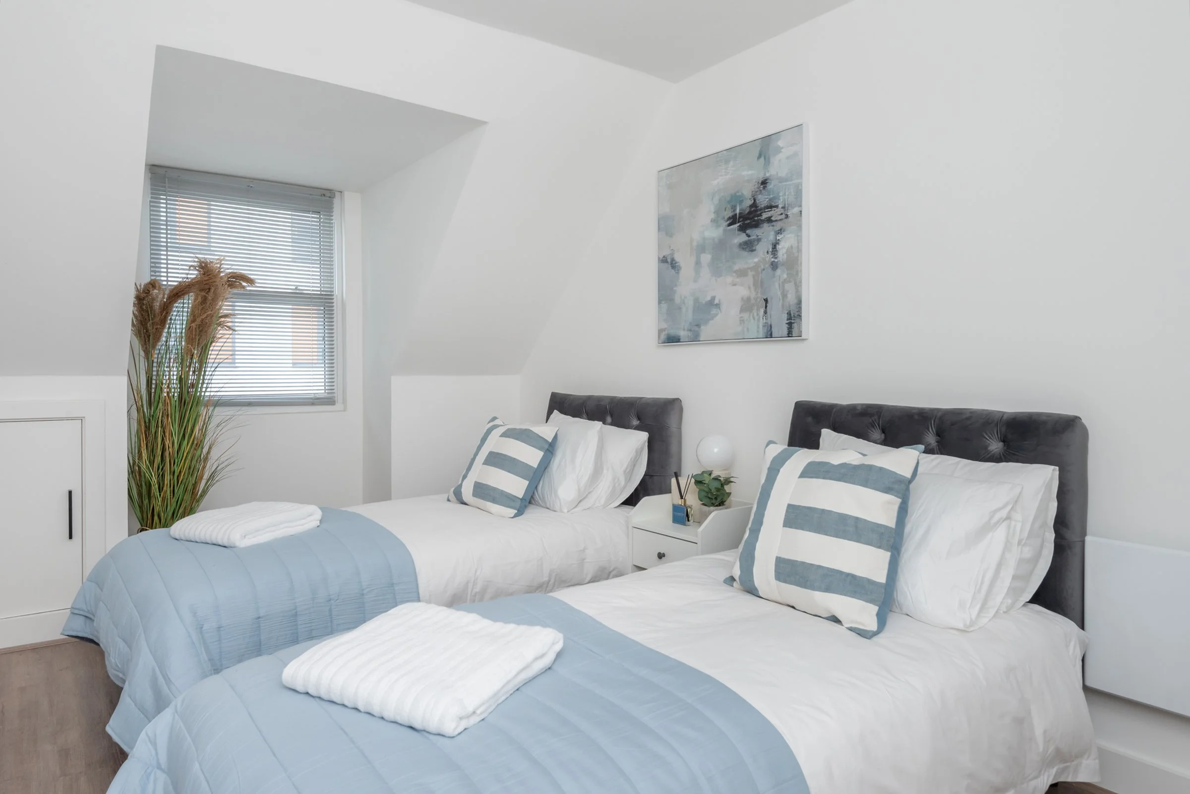 A bedroom with two single beds with gray tufted headboards, white bedding, and light blue quilts. Each bed has a striped pillow, and towels are folded at the foot of each bed. There is a small white nightstand between the beds with a modern lamp, a small plant, and a decorative item. A window with blinds is on the left, and a large abstract painting is on the wall above the beds. A tall decorative plant with dried grasses is positioned in the corner near the window.