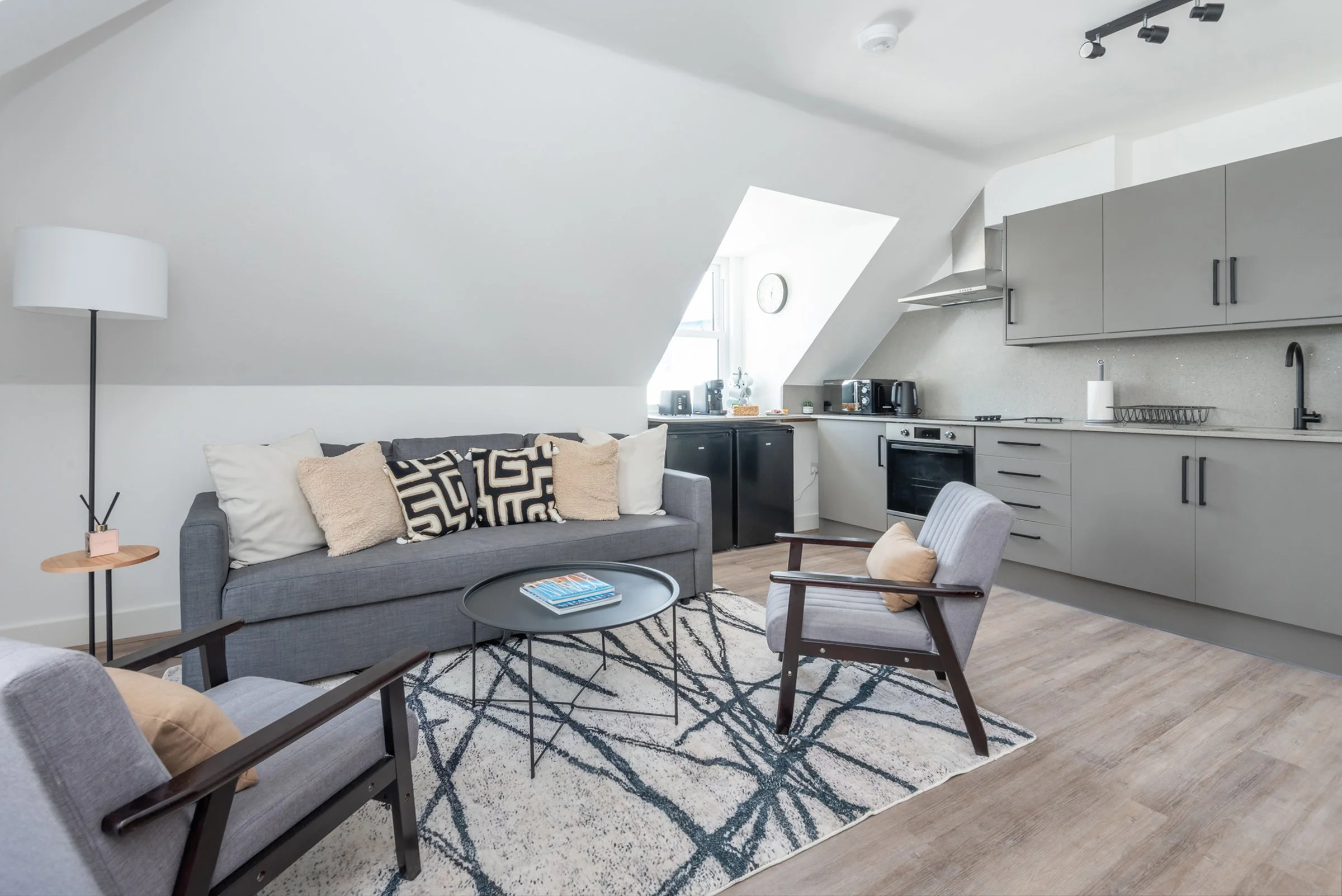 Living room with gray sofa, armchairs, black and white patterned pillows, coffee table on a geometric rug, and a compact kitchen with gray cabinets, countertop, and appliances under a sloped ceiling with a skylight.