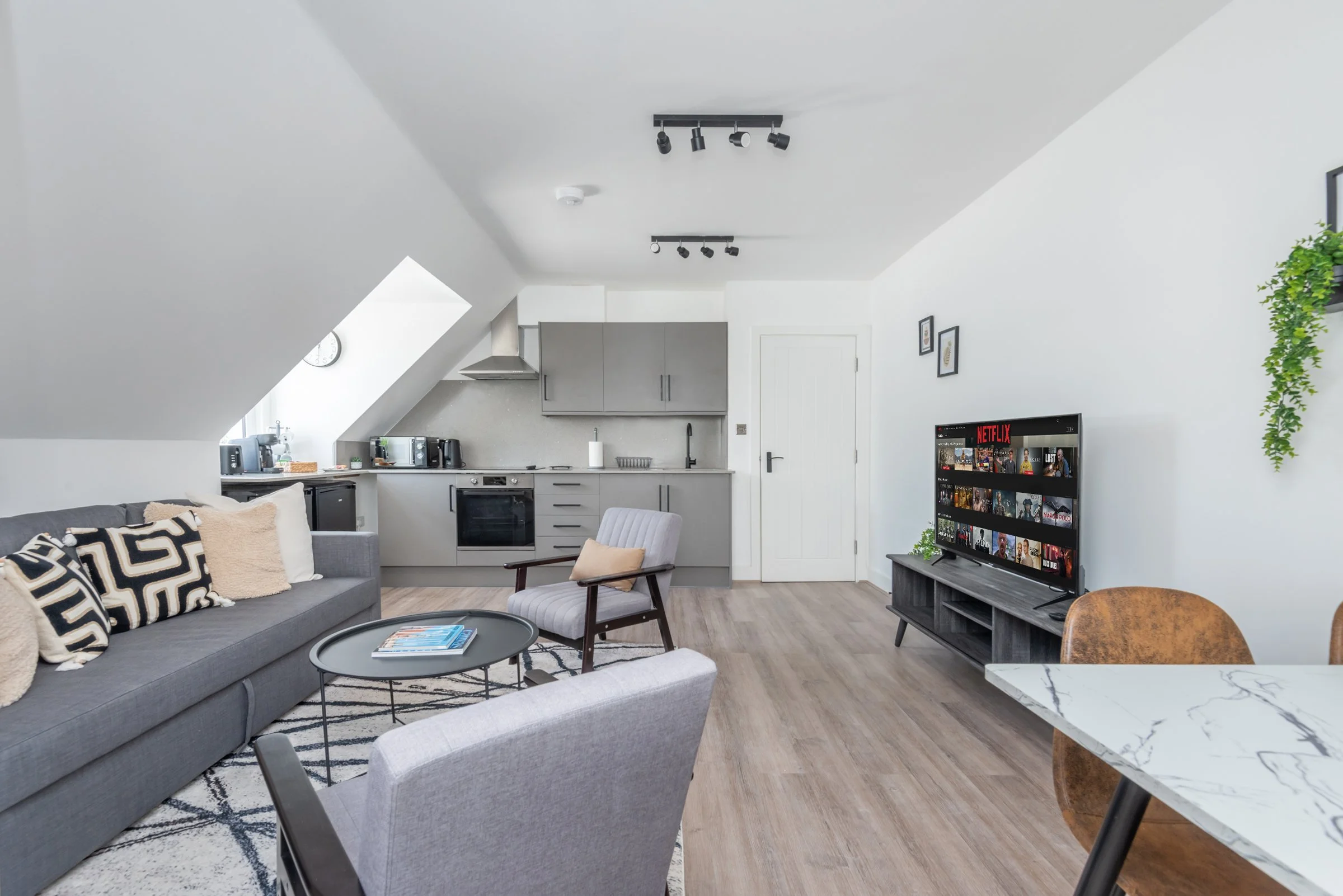 Living room with a grey sofa, two accent chairs, a round coffee table, and a TV on a stand. The background shows a small kitchen with grey cabinets and appliances, and the wall is decorated with framed artwork and a hanging plant.