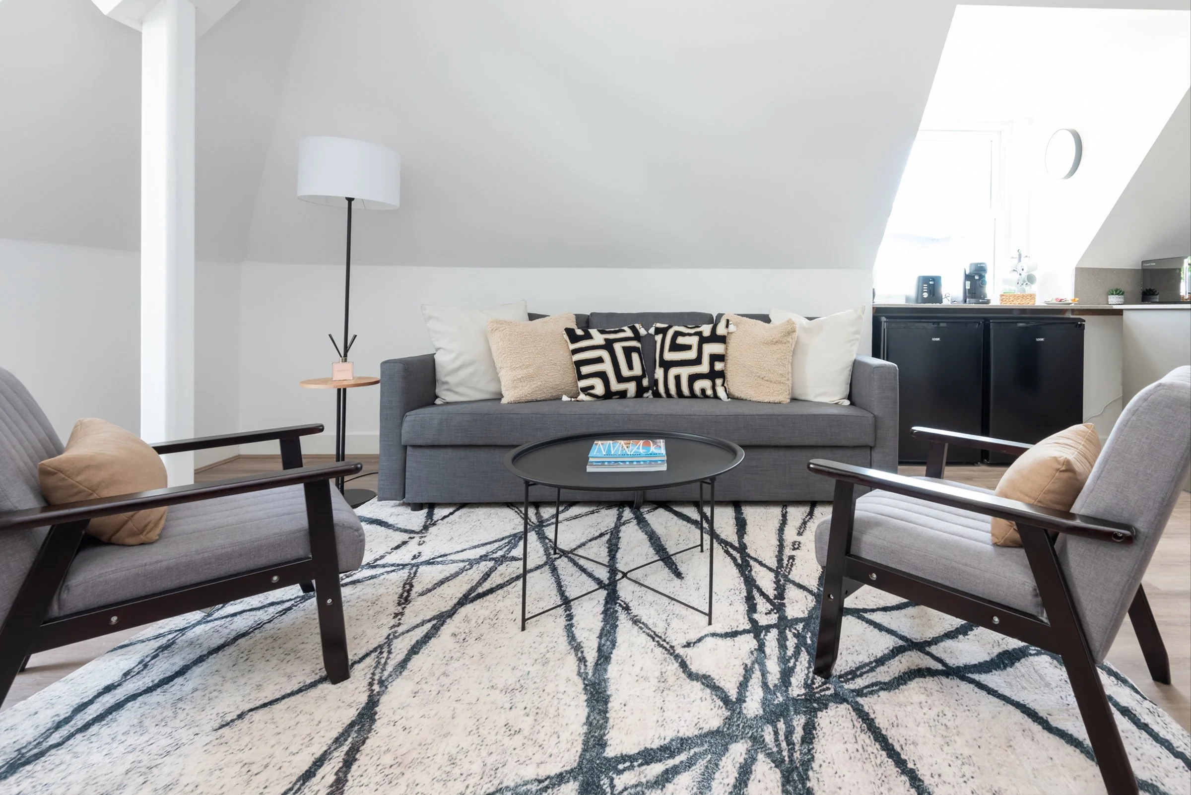 Living room with gray sofa, two gray armchairs, a black round coffee table, and a patterned area rug. A white floor lamp is next to the sofa. Small black cabinets and windows are in the background.