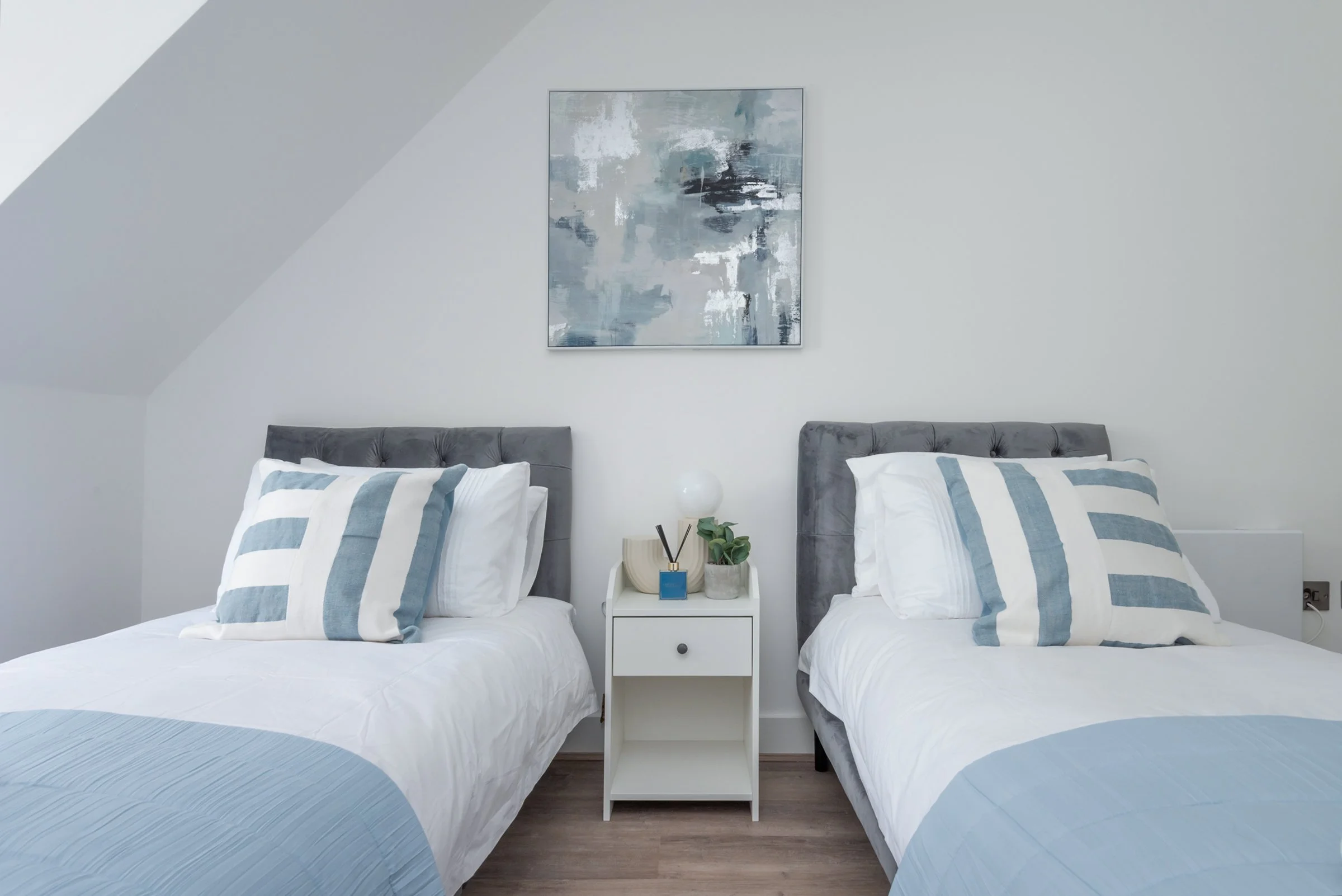 Two neatly made twin beds with gray upholstered headboards in a minimalistic bedroom, each with white and blue striped pillows and bedspreads, a white nightstand with a lamp, decorative item, and green plant between the beds, and an abstract painting hanging on the white wall.