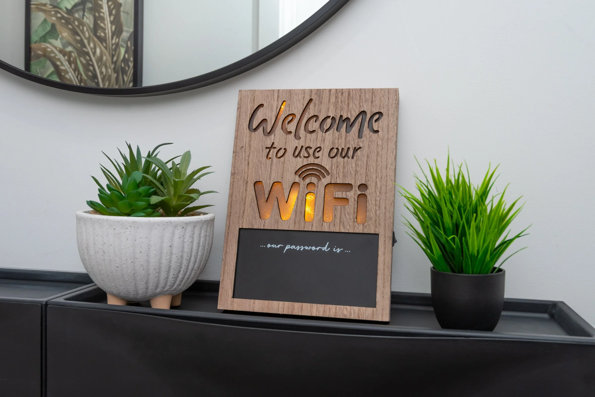 Wooden sign with 'Welcome to use our WiFi' illuminated letters and a black chalkboard space with '...our password is...' text, flanked by potted succulents on a black reception desk, with a mirror and framed plant picture reflected in the background mirror.