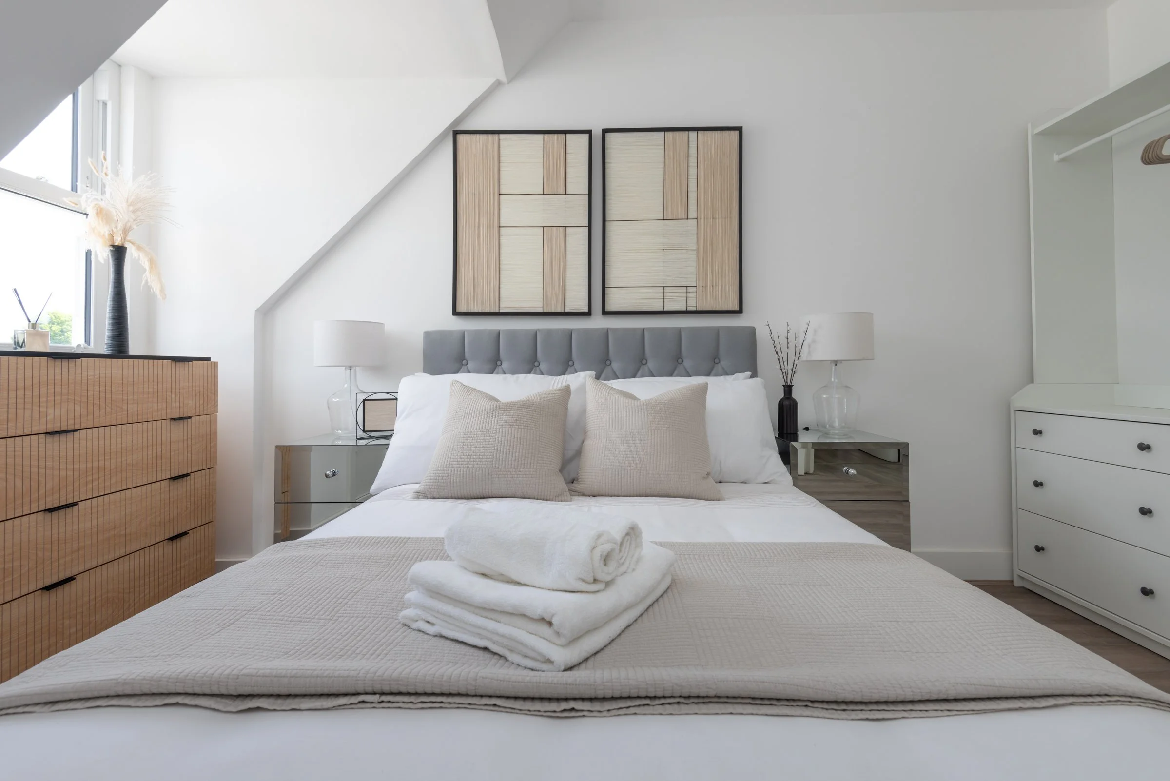 Modern bedroom with a neatly made bed, beige pillows, and a folded towel on top. The room has white walls, a gray upholstered headboard, and minimalist furniture, including a wooden dresser on the left and white drawers on the right. Two framed abstract art pieces hang above the bed, and natural light enters through a window on the left.