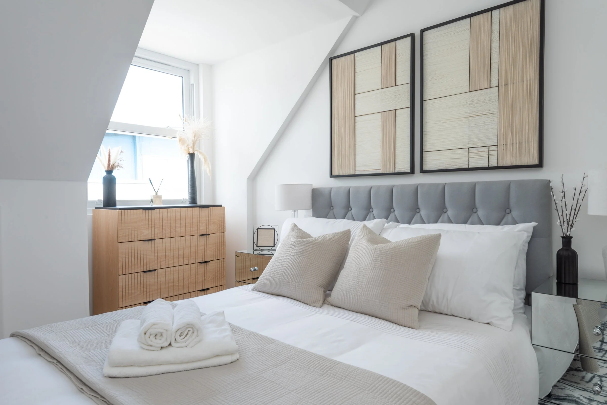 A modern, minimalist bedroom with a bed, beige pillows, rolled towels on the bed, a wooden dresser, decorative vases, and wall art.