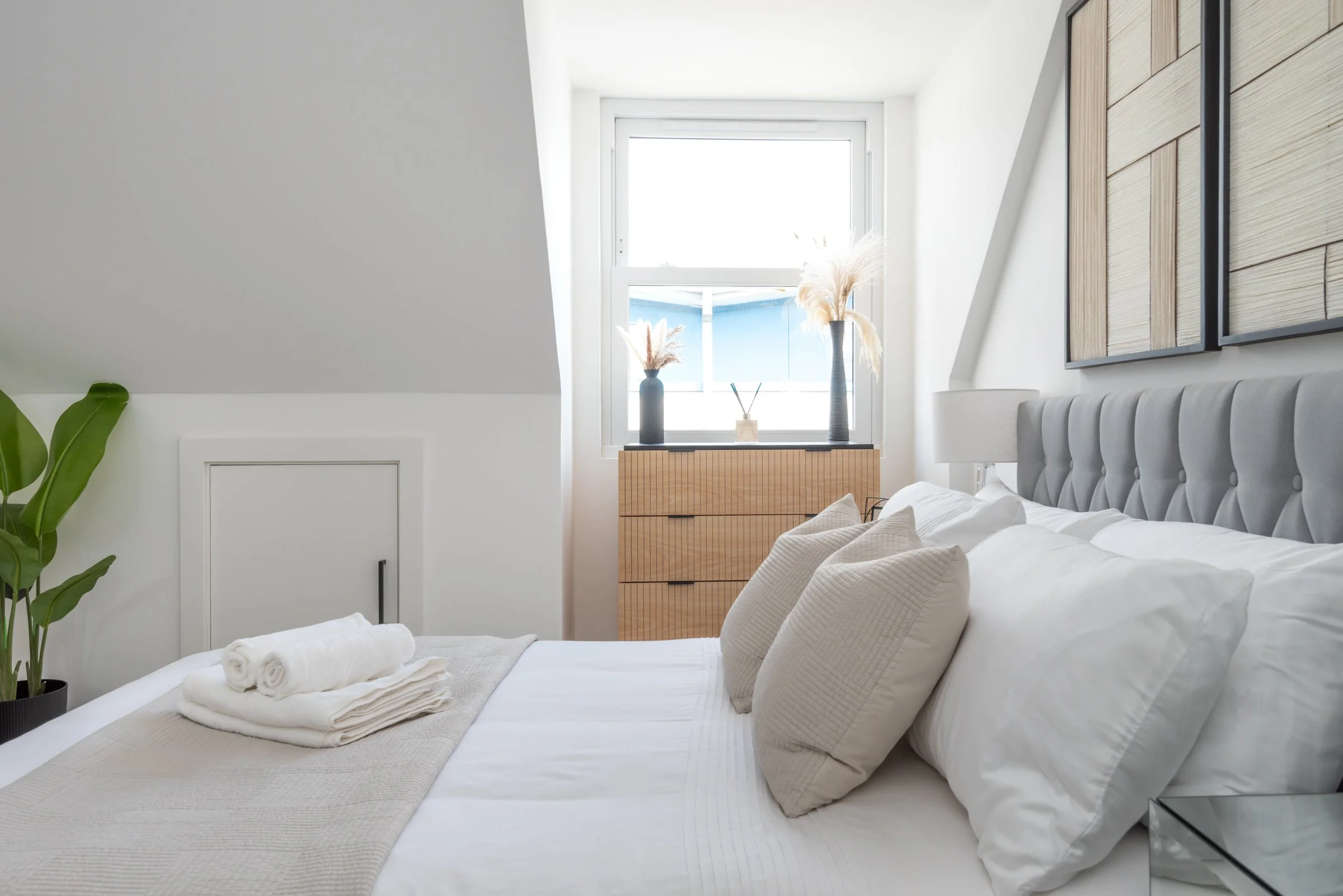 A neatly made bed with white bedding and beige pillows in a bright, minimalistic bedroom with a window, a wooden dresser, and decorative vases with pampas grass.