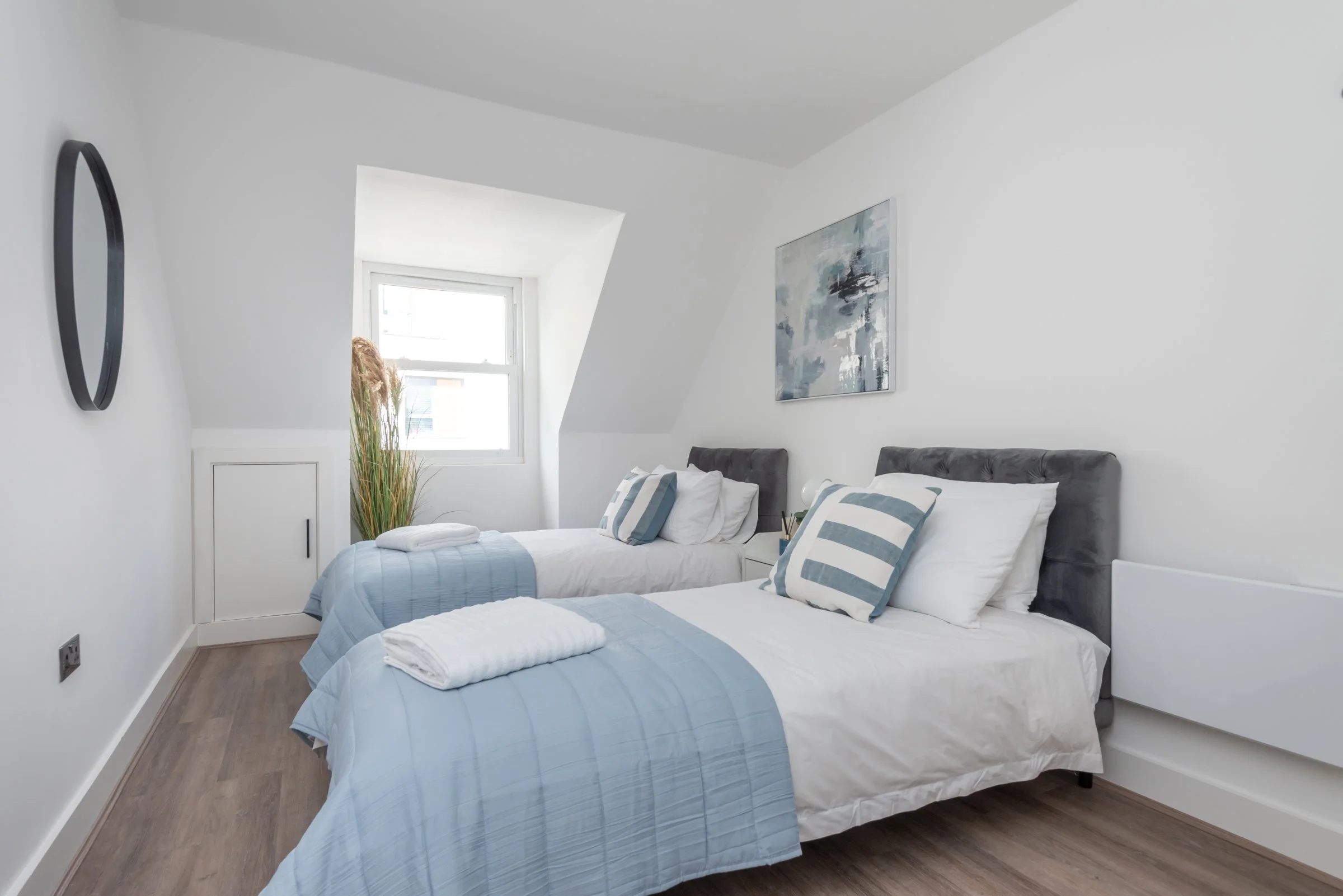 A bright, minimalistic bedroom with two single beds, each with a grey headboard, white bedding, and blue striped pillows. There's a small nightstand between the beds, a window on the back wall, a round mirror on the left wall, and an abstract painting above the beds.