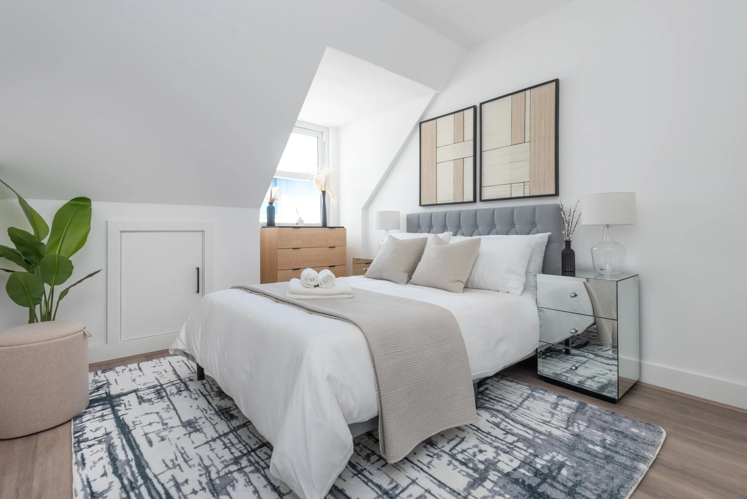 Modern bedroom with white walls, gray upholstered headboard, a mirrored nightstand, wooden dresser, framed artwork, potted plant, and a window with natural light