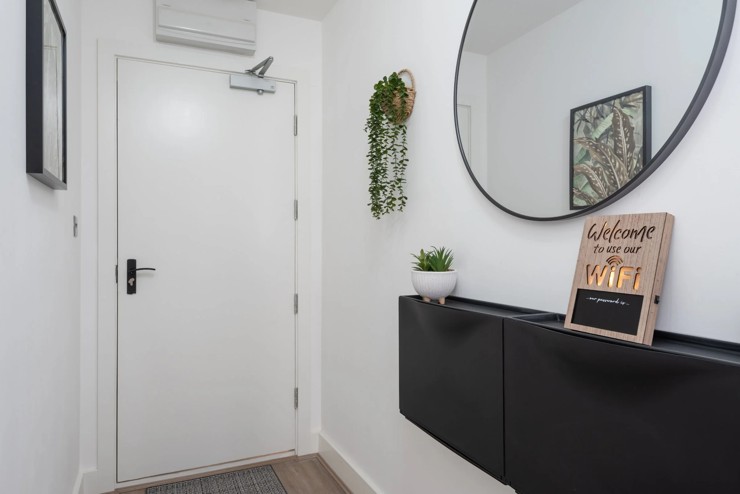 Modern entryway with white walls, a white door, and a black wall-mounted cabinet. Decor includes a round mirror, a framed botanical print, a hanging potted plant, a small potted succulent, and a sign indicating WiFi password.