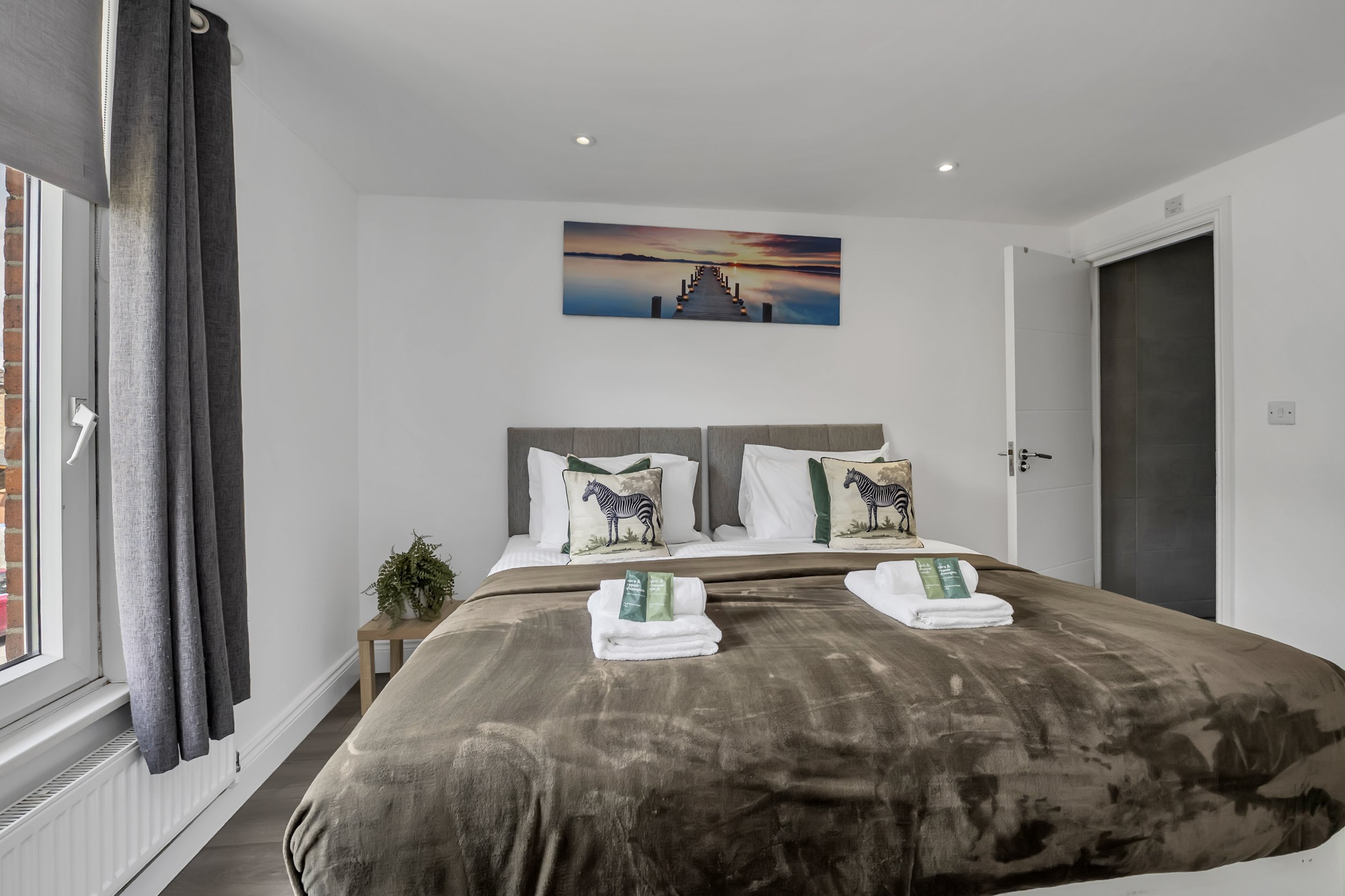 A neatly made bed with two pillows that have zebra and jungle animal designs, two white towels with green packaging, and a brown bedspread in a bedroom with white walls, a landscape painting of a pier at sunset above the bed, a window with gray curtains, and a small bedside plant.