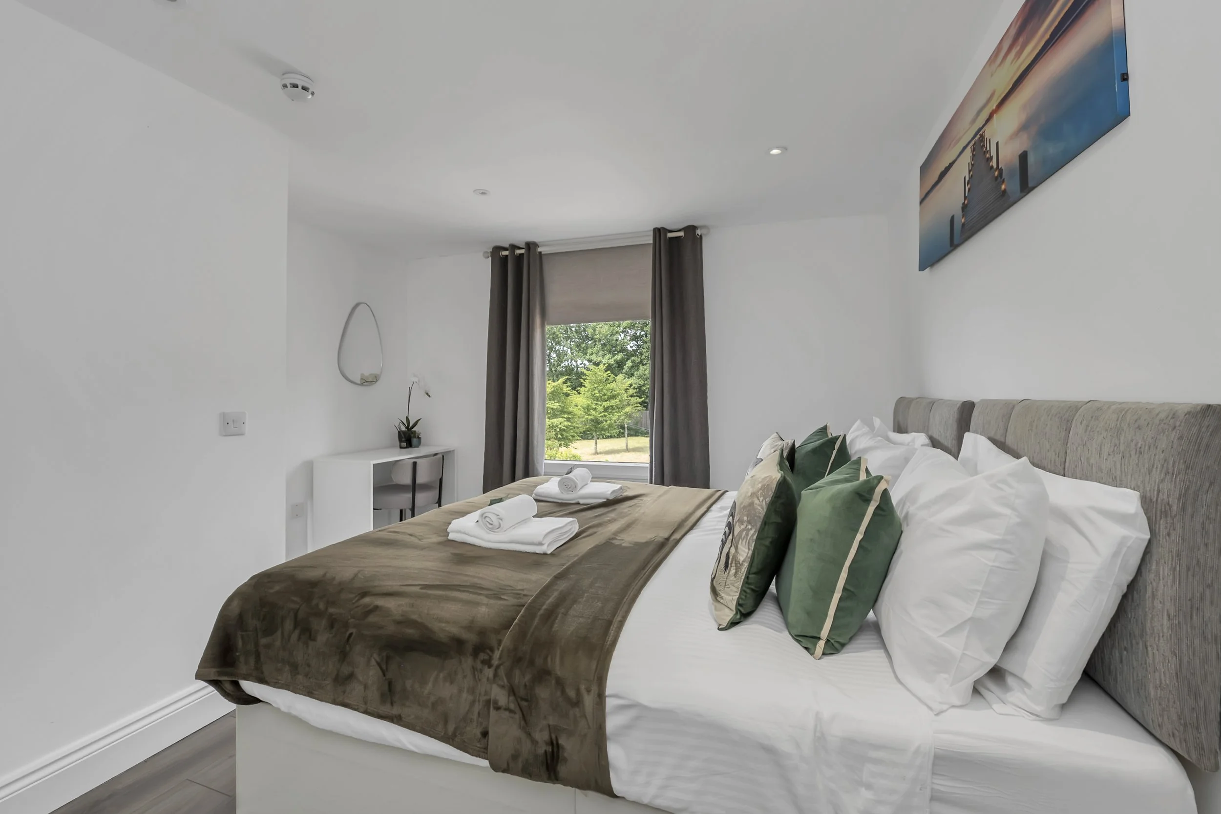 A neatly made bedroom with a large bed, white sheets, multiple pillows, a brown throw blanket, and two rolled white towels on the bed. There is a window with brown curtains, a small white desk with a mirror, and a painting of a pier at sunset on the wall.