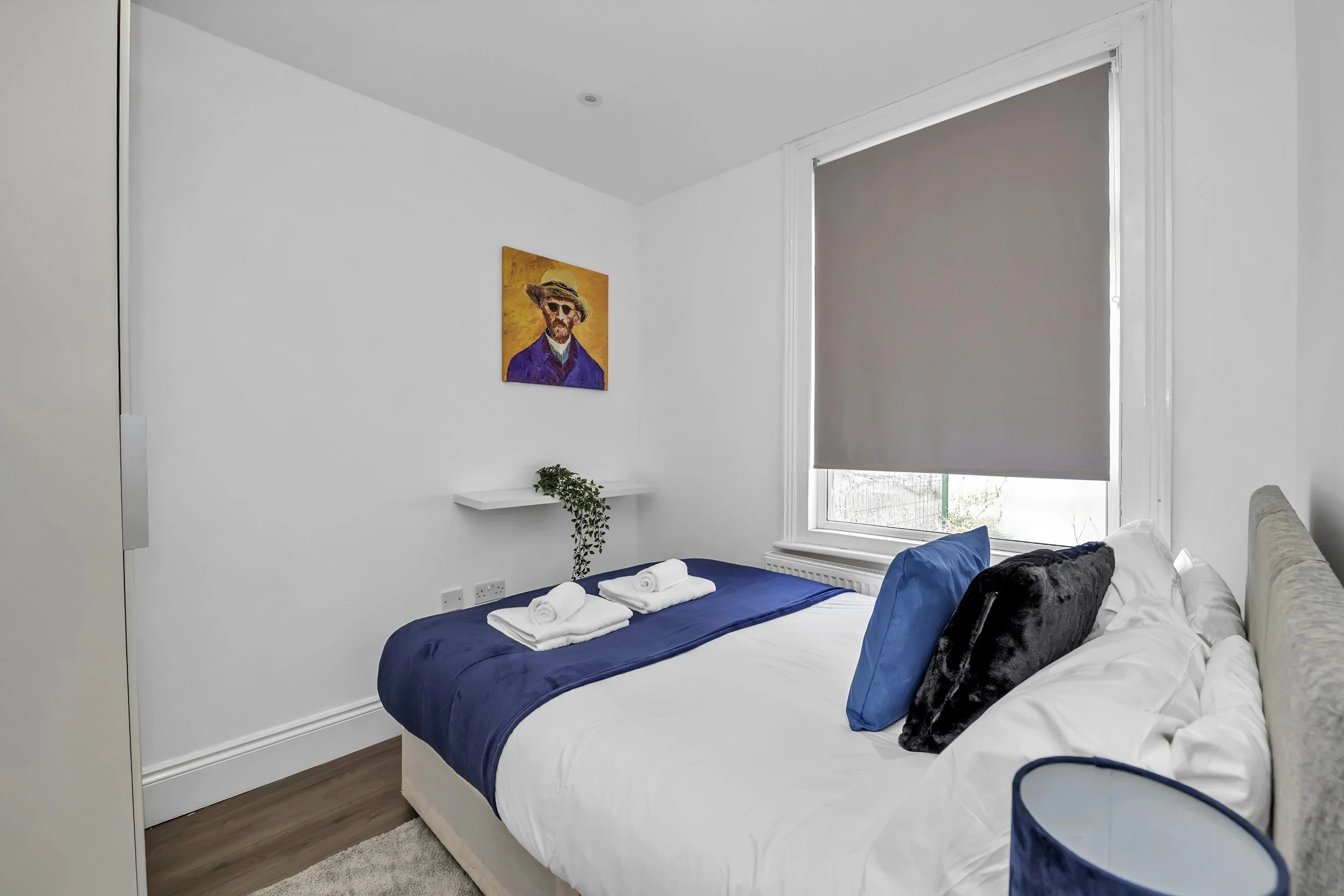 A neatly made bed with blue and white bedding, two rolled towels on top, two pillows, and a black cushion. A window with closed gray blinds, a small shelf with a plant, a colorful portrait on the wall, and a bedside table with a blue lampshade are visible in a bright, minimalist bedroom.