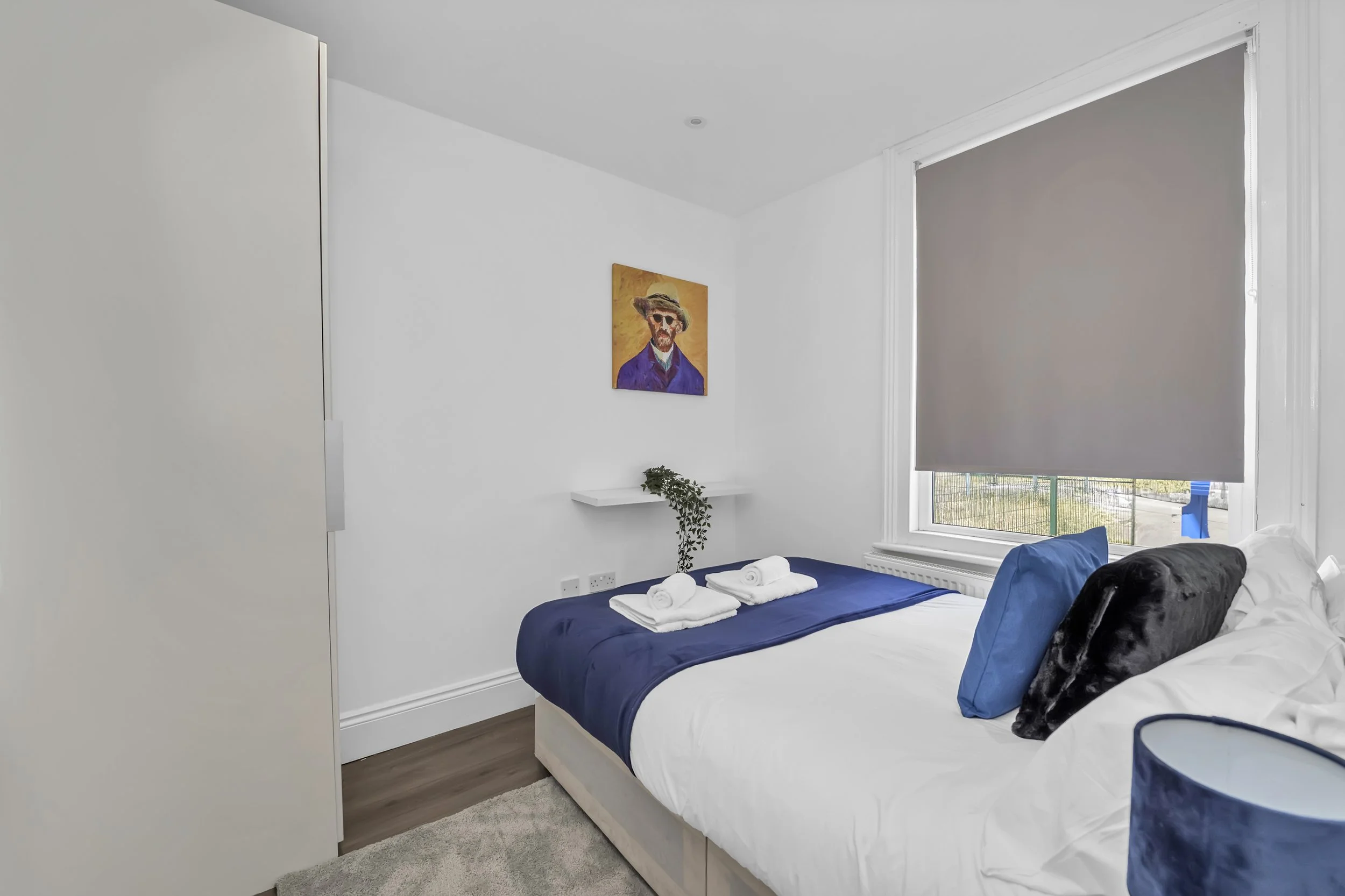 A neatly made bed with white sheets, blue blanket, and pillows in a minimalist bedroom. A window with a closed gray blind and a small wall shelf with a potted plant above the bed. A colorful portrait hangs on the white wall.