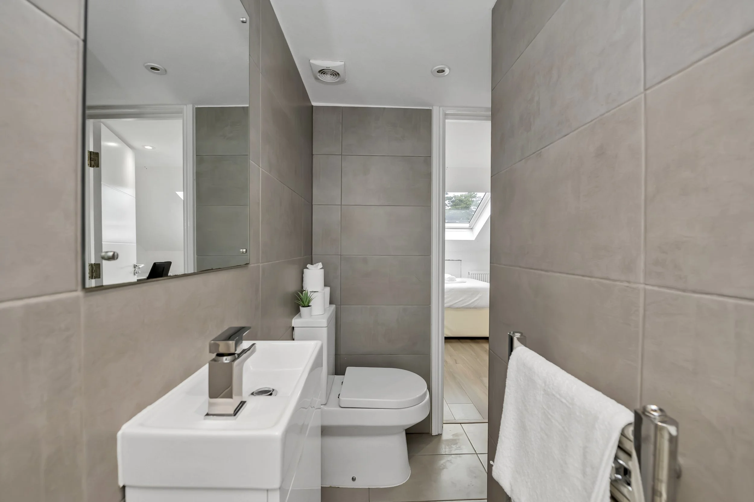 Small modern bathroom with beige tiled walls, white sink, toilet with a closed lid, mirror, small decorative plant, and a white towel on a silver towel rack. Through the doorway, a bedroom with a bed and a skylight window is visible.