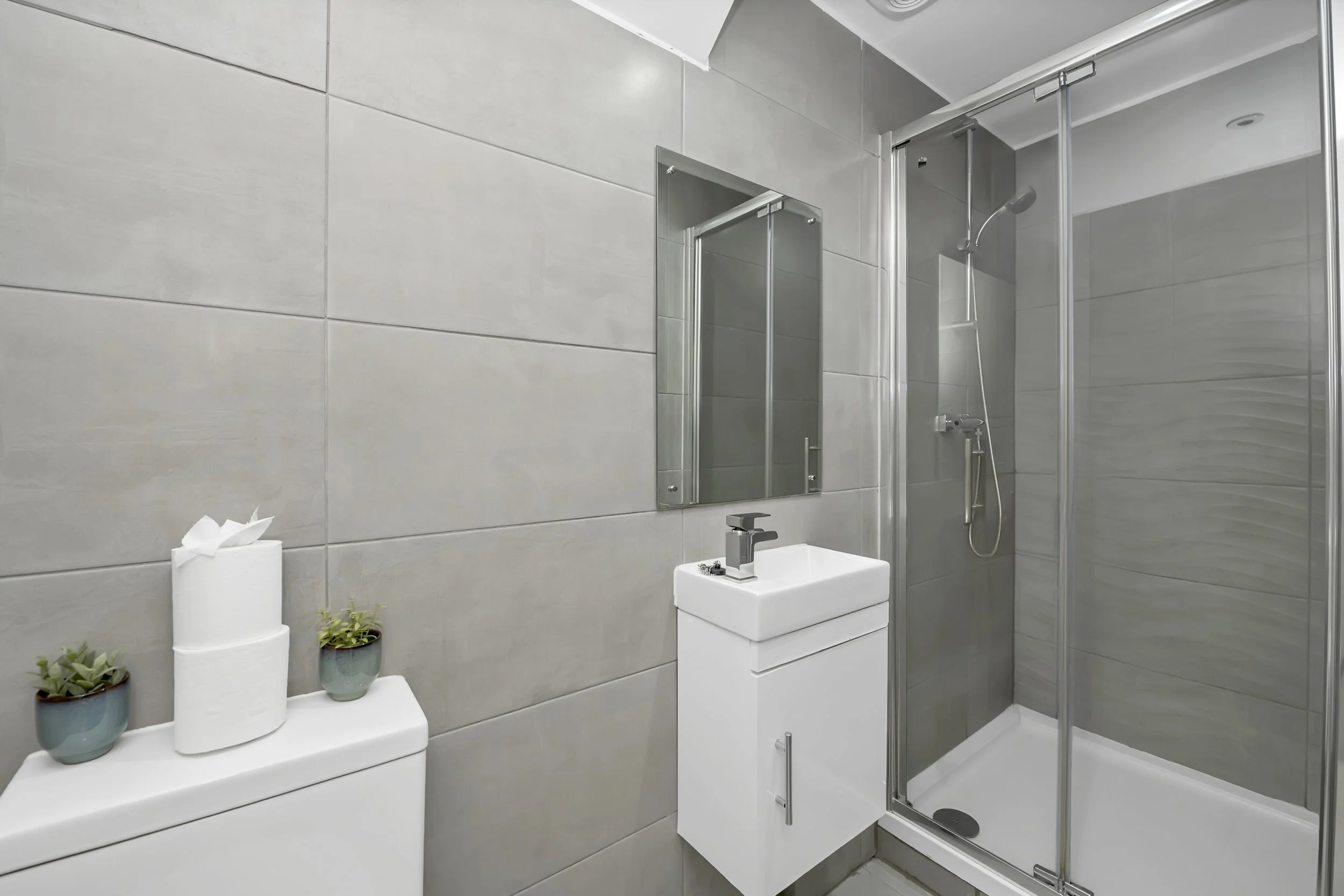 A modern bathroom with gray tiled walls, a compact white sink with a black faucet, a mirror above the sink, a glass-enclosed shower with a showerhead, and a white toilet topped with two small potted plants and a roll of toilet paper.