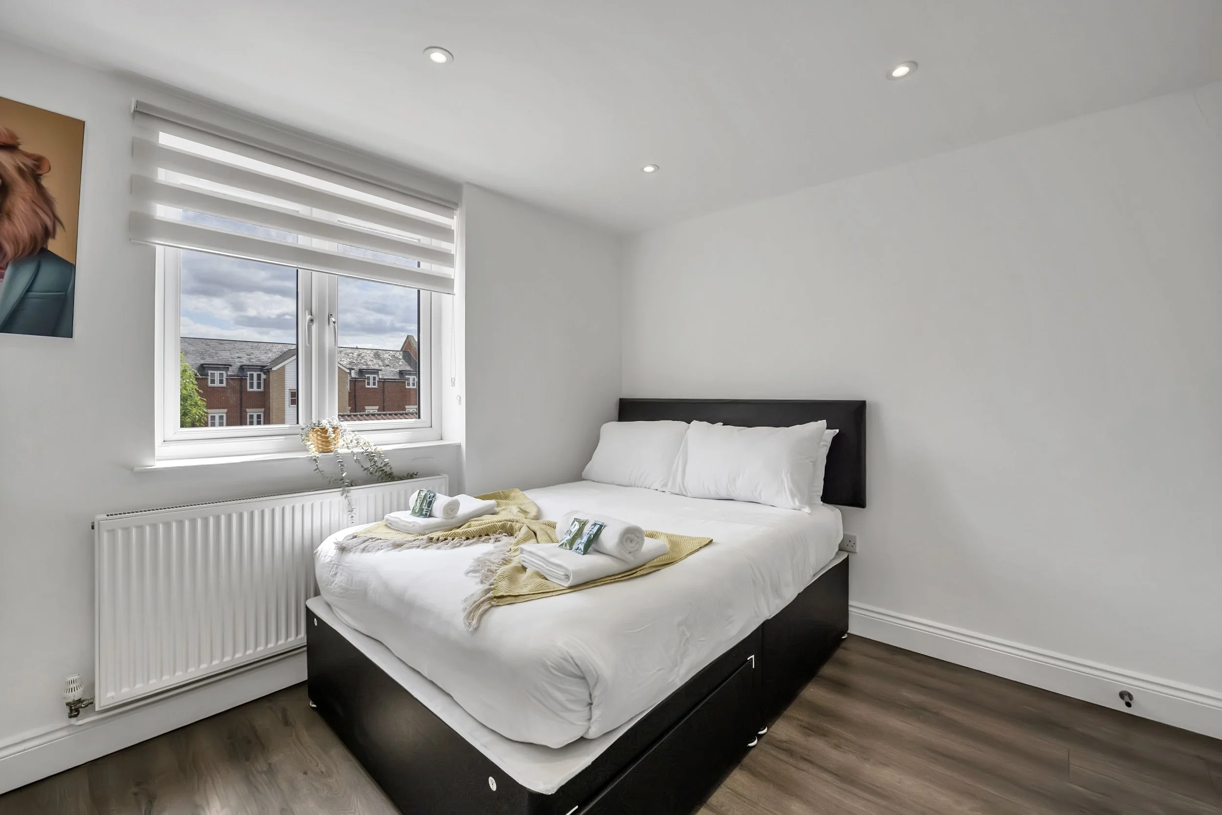 A clean, modern bedroom with a bed dressed in white linens and pillows, towels with small bottles on top, a window with daytime view of residential buildings, painted white walls, and a wooden floor.