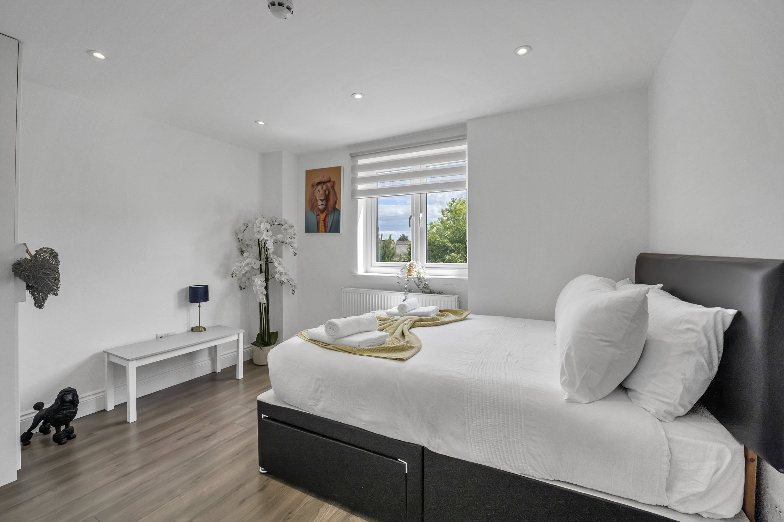 A modern bedroom with a white bed, pillows, and linens. There are rolled towels on the bed. The room has a window with blinds, a small bedside table with a blue lamp, a wall art of a lion, a tall decorative plant, and some wall decorations. The floor is wood, and the walls are white.
