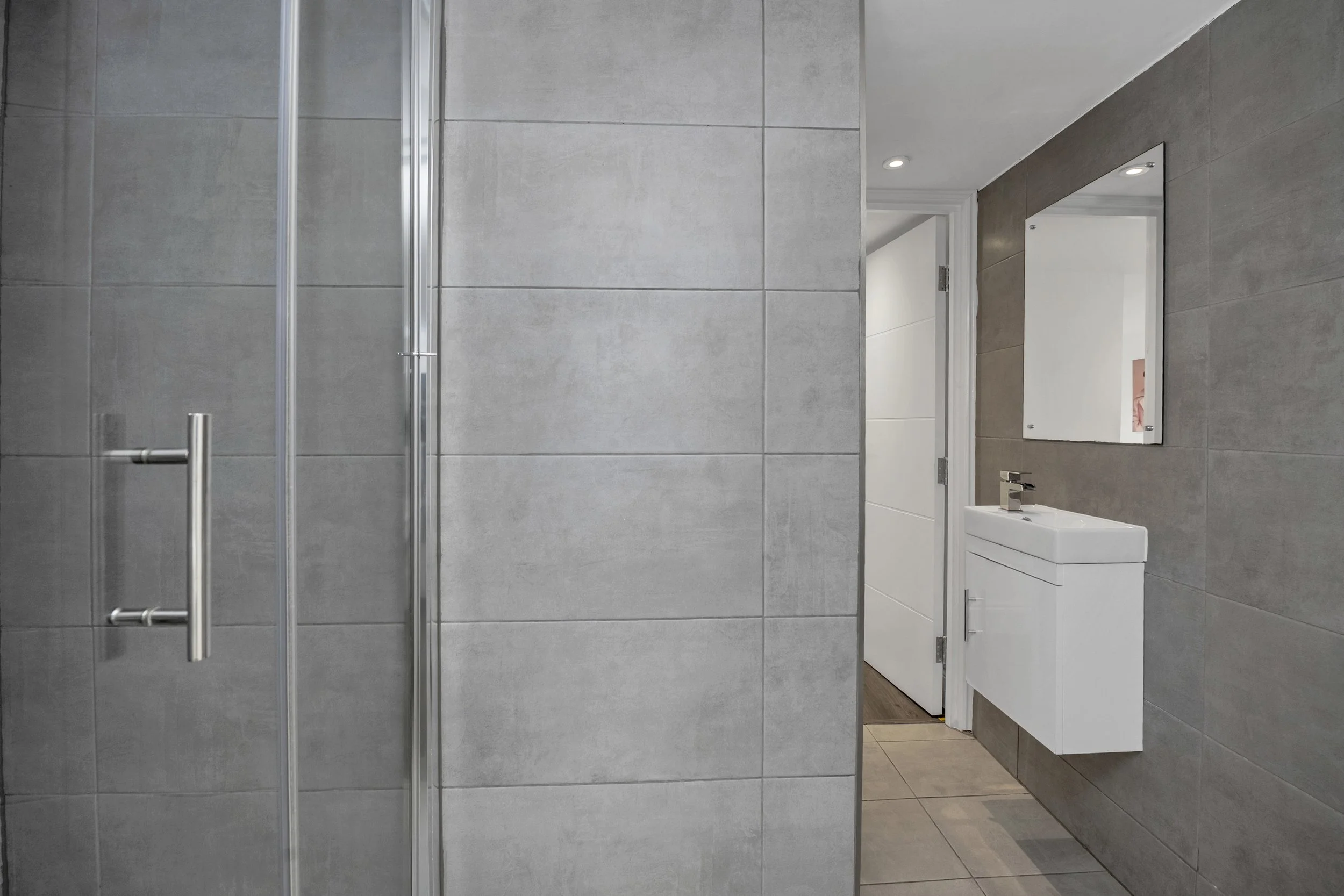 Modern bathroom with gray tiled walls, a glass shower door on the left, a white floating sink with a mirror on the right, and a doorway leading to another room.