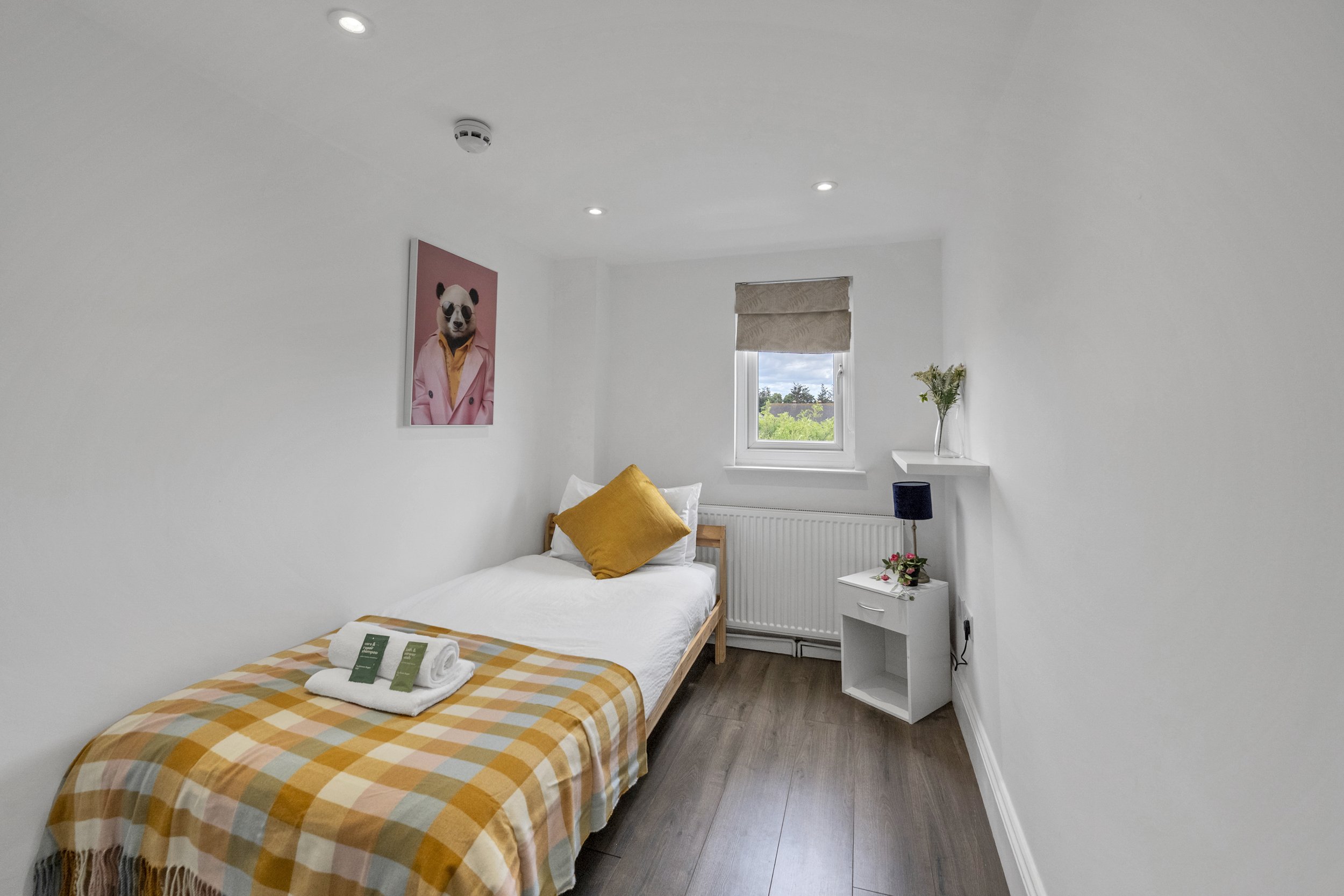 A small bedroom with white walls, a single bed with white bedding and a patterned blanket, a gold pillow, and towels on the bed. There is a window with a beige shade, a side table with a small lamp and flower arrangement, and a wall art of a panda in a pink jacket.