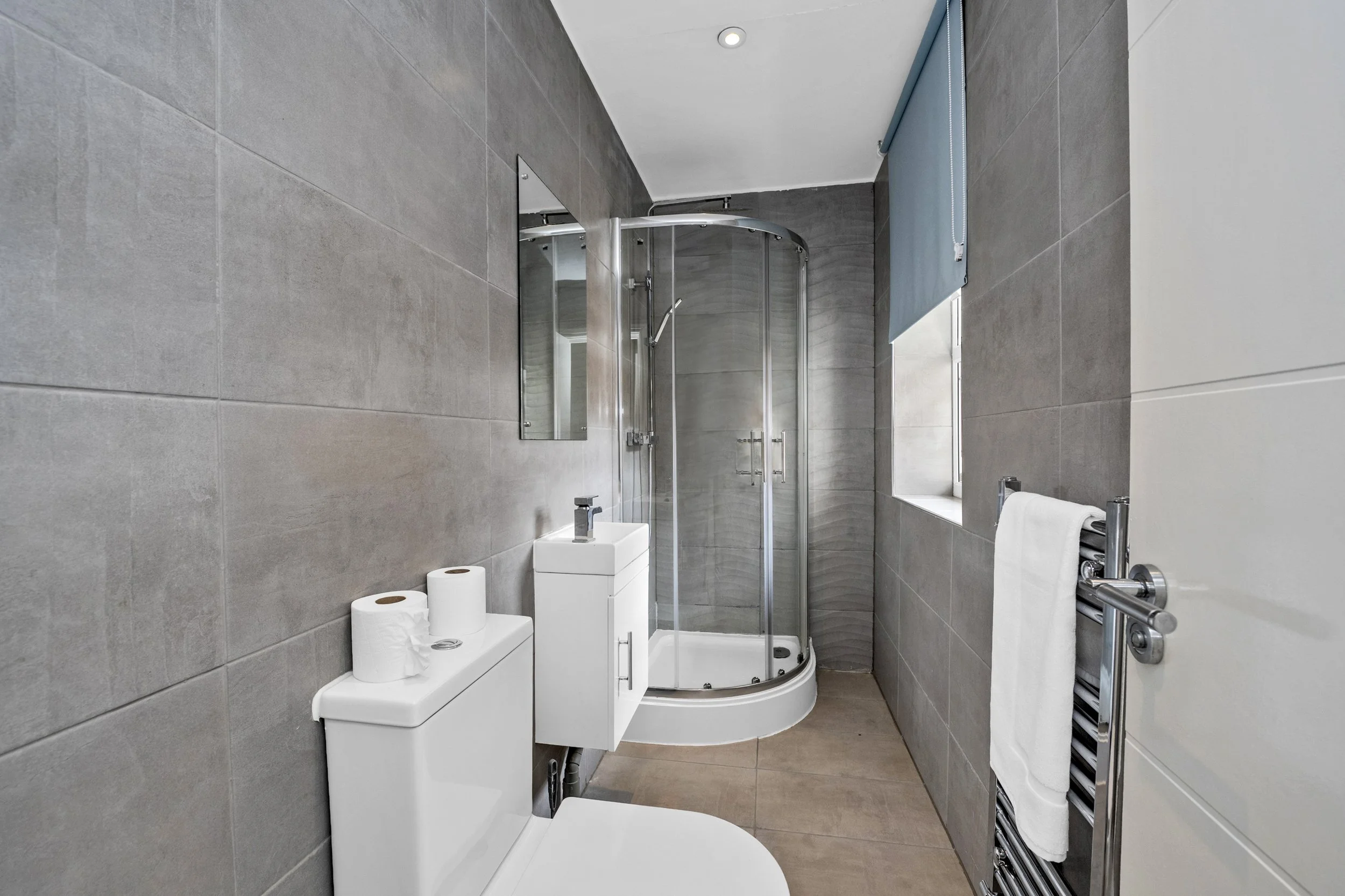 Modern bathroom with gray tiled walls, a corner shower stall with glass doors, a white toilet, a small white sink, a mirror, a window with a blue roller blind, and a heated towel rail with a white towel.