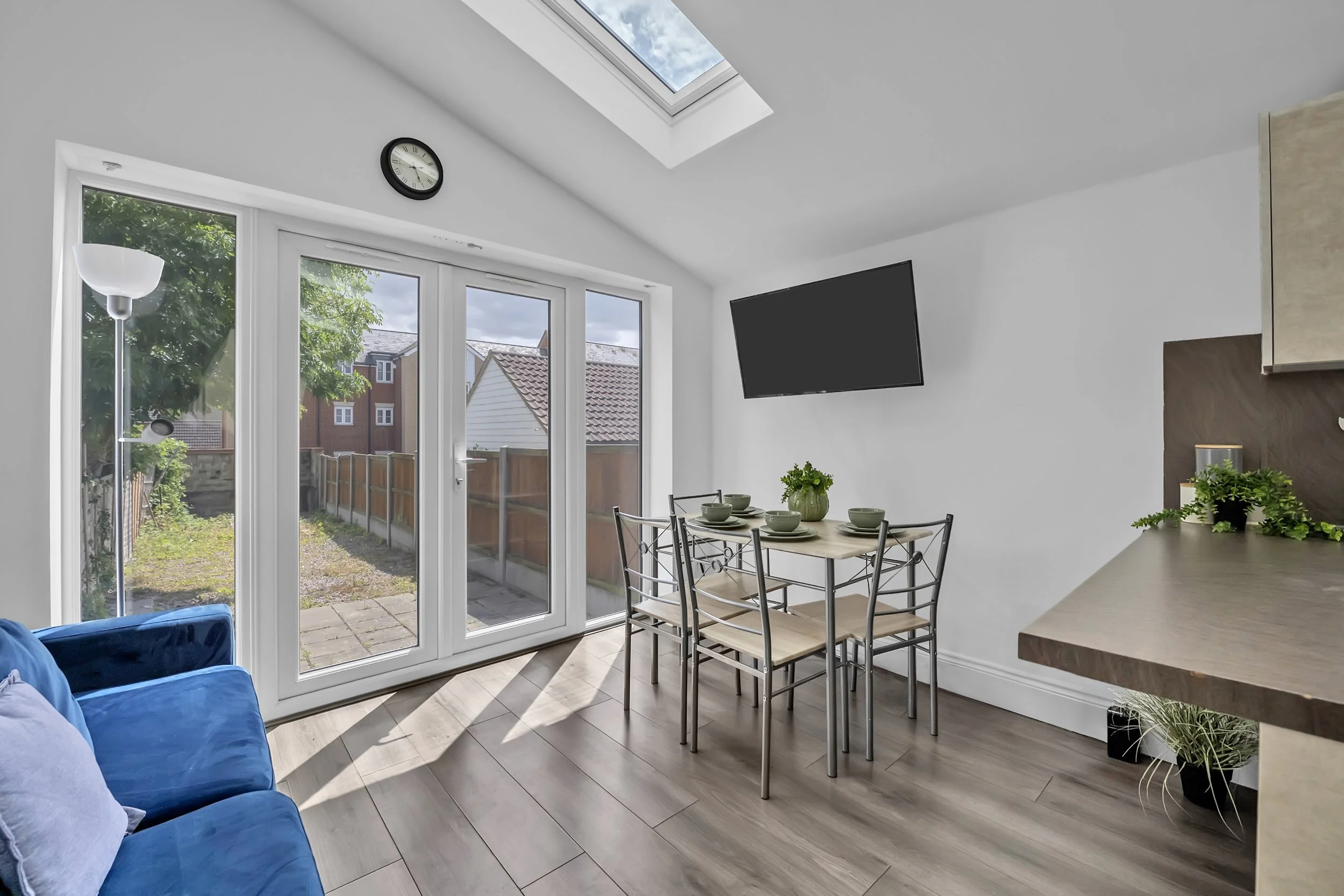 A bright living room with large glass doors leading to a backyard, a blue sofa on the left, a wooden dining table with chairs in the center, a wall-mounted TV, a wall clock, and a skylight letting in natural light.