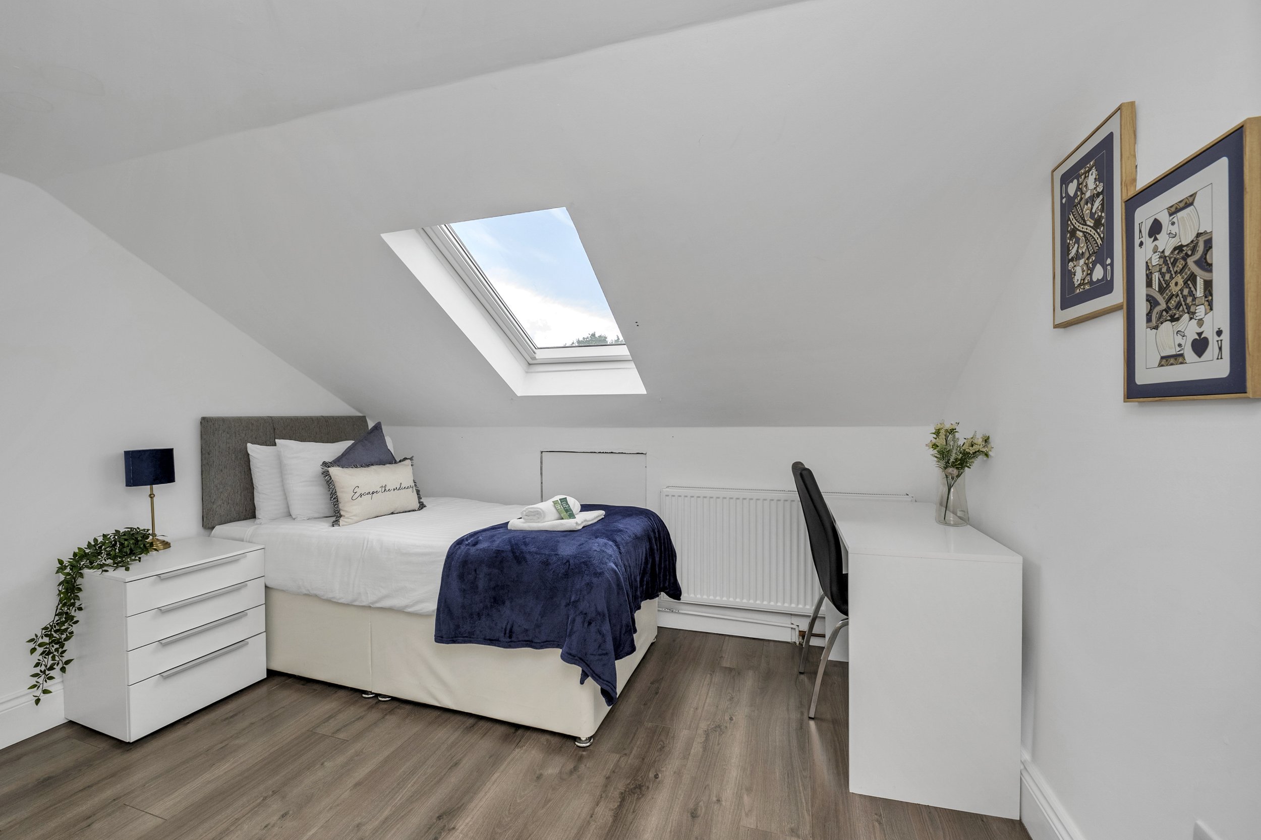 A cozy bedroom in an attic with white walls and wood flooring. Features a bed with white linens, a navy blue blanket, and pillows, including one with the phrase "Escape the ordinary". A skylight window shows a blue sky with clouds. A white nightstand with a trailing plant, a black table lamp, and a small desk with a black chair. Two framed artistic prints hang on the wall.