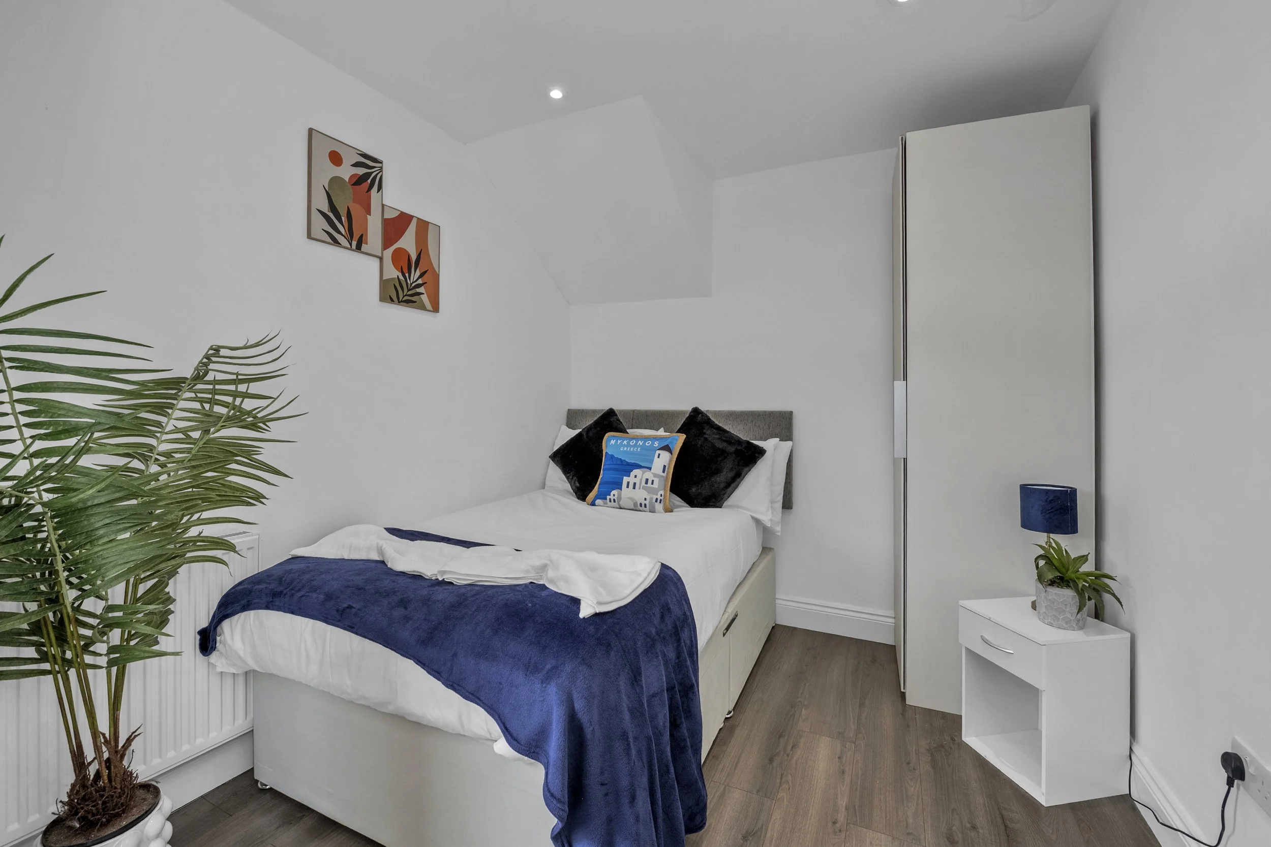 Small bedroom with white walls, a bed with a grey headboard, white bedding, navy blue blanket, and decorative pillows, including a Greek-themed cushion. There is a white nightstand with a potted plant and a blue lamp, a wardrobe, a tall green plant, and framed abstract art on the wall.