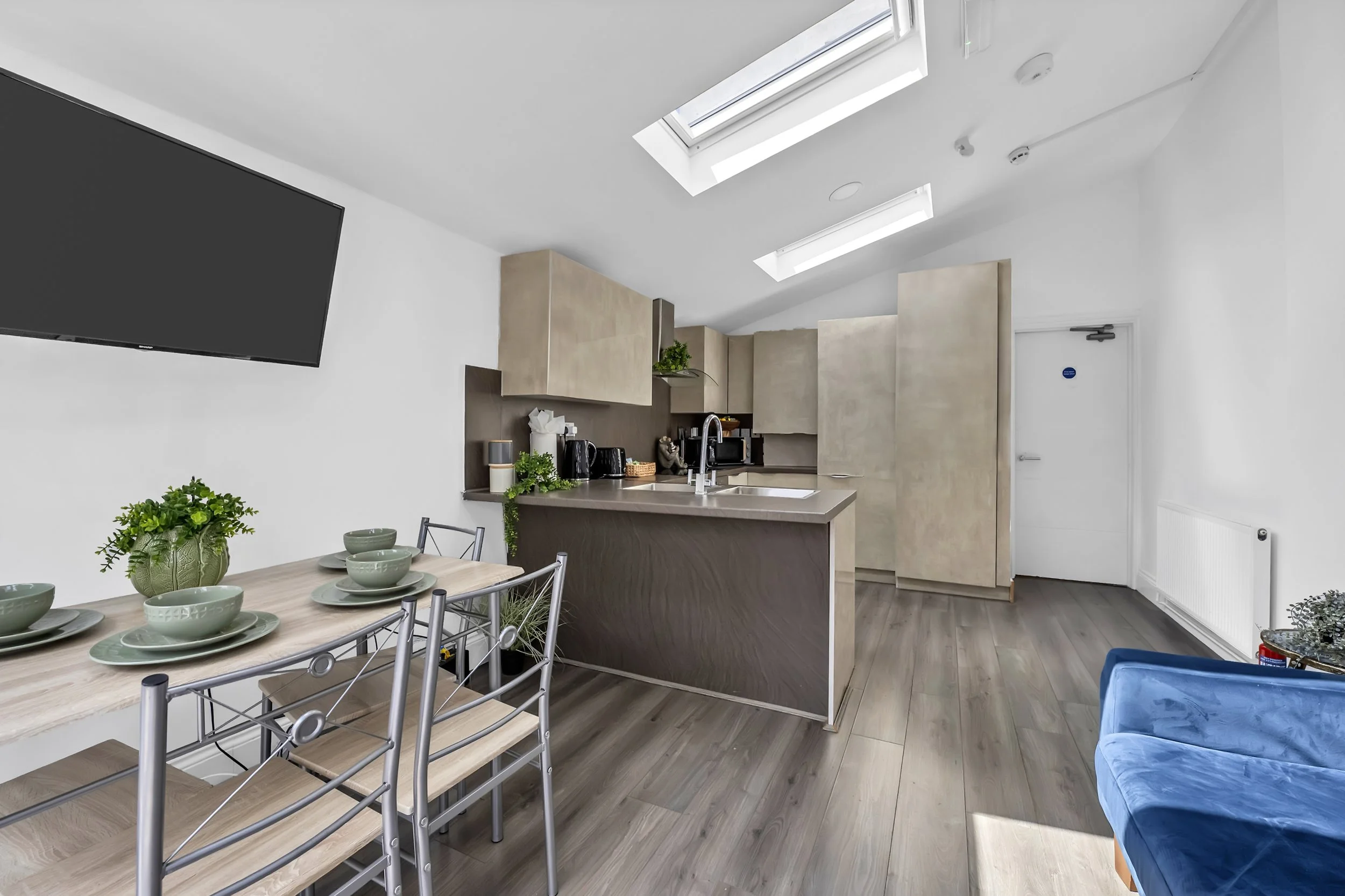 Modern kitchen and dining area with skylights, beige cabinets, a kitchen island with a sink, a wall-mounted TV, a dining table with six chairs, a blue armchair, and wood flooring.