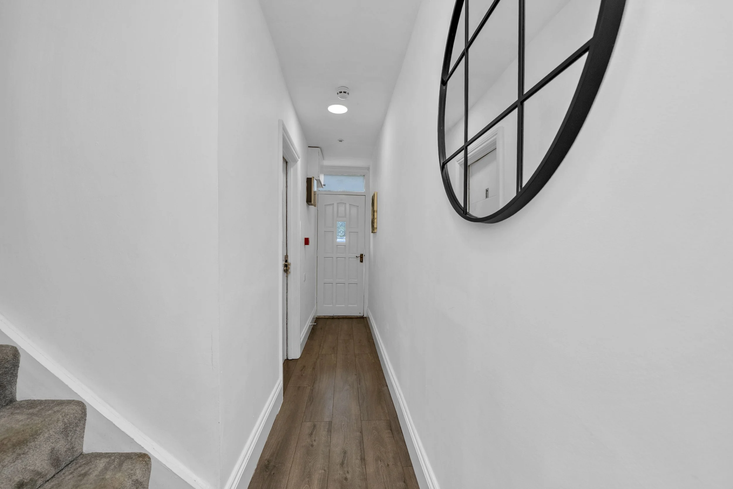 Empty hallway inside a house with white walls, wooden flooring, a large round black wall clock, and a door at the end with a window above.